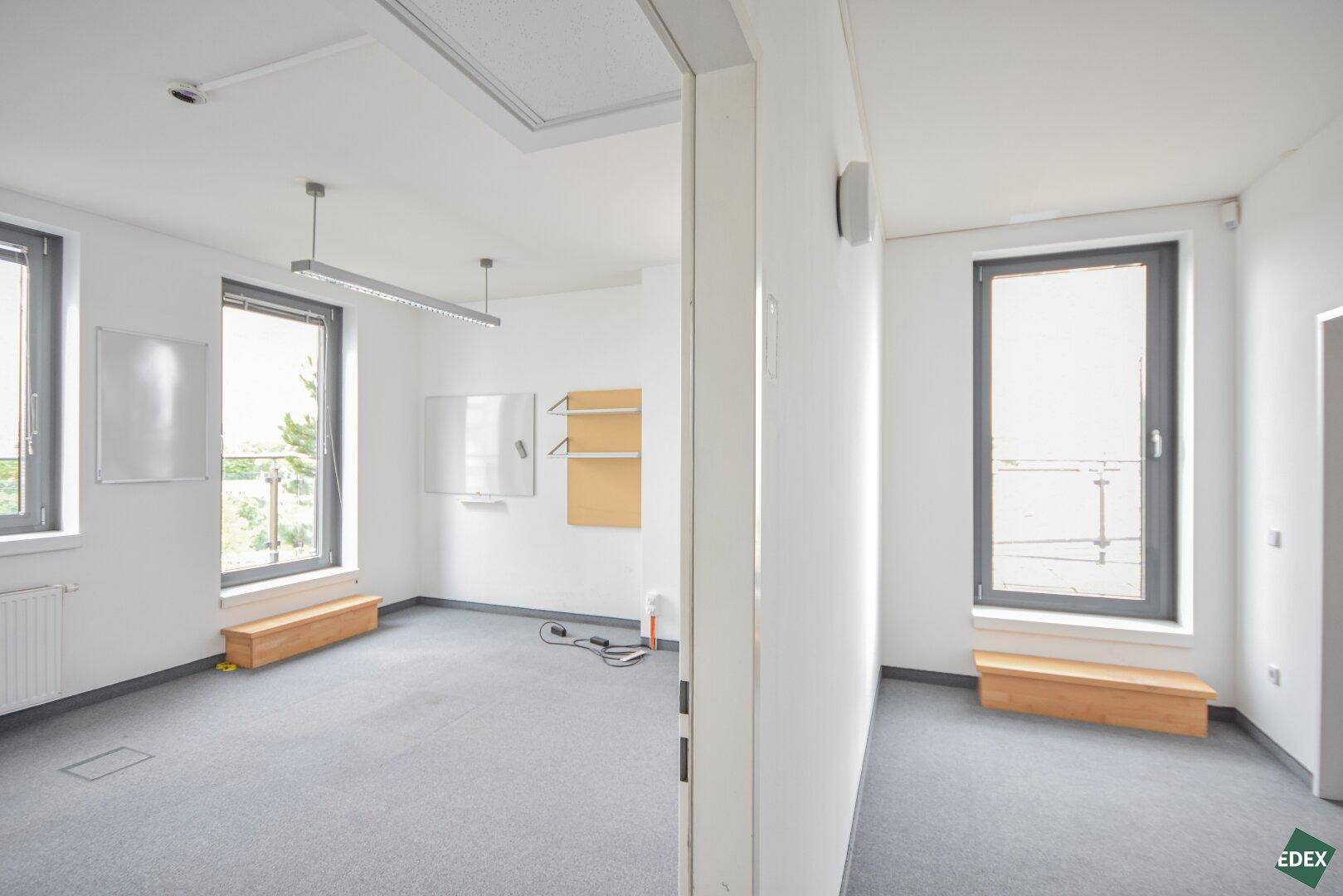 Helles Büro mit Teppichboden, großen Fenstern und einfacher Ausstattung, ideal für Arbeitsplätze.