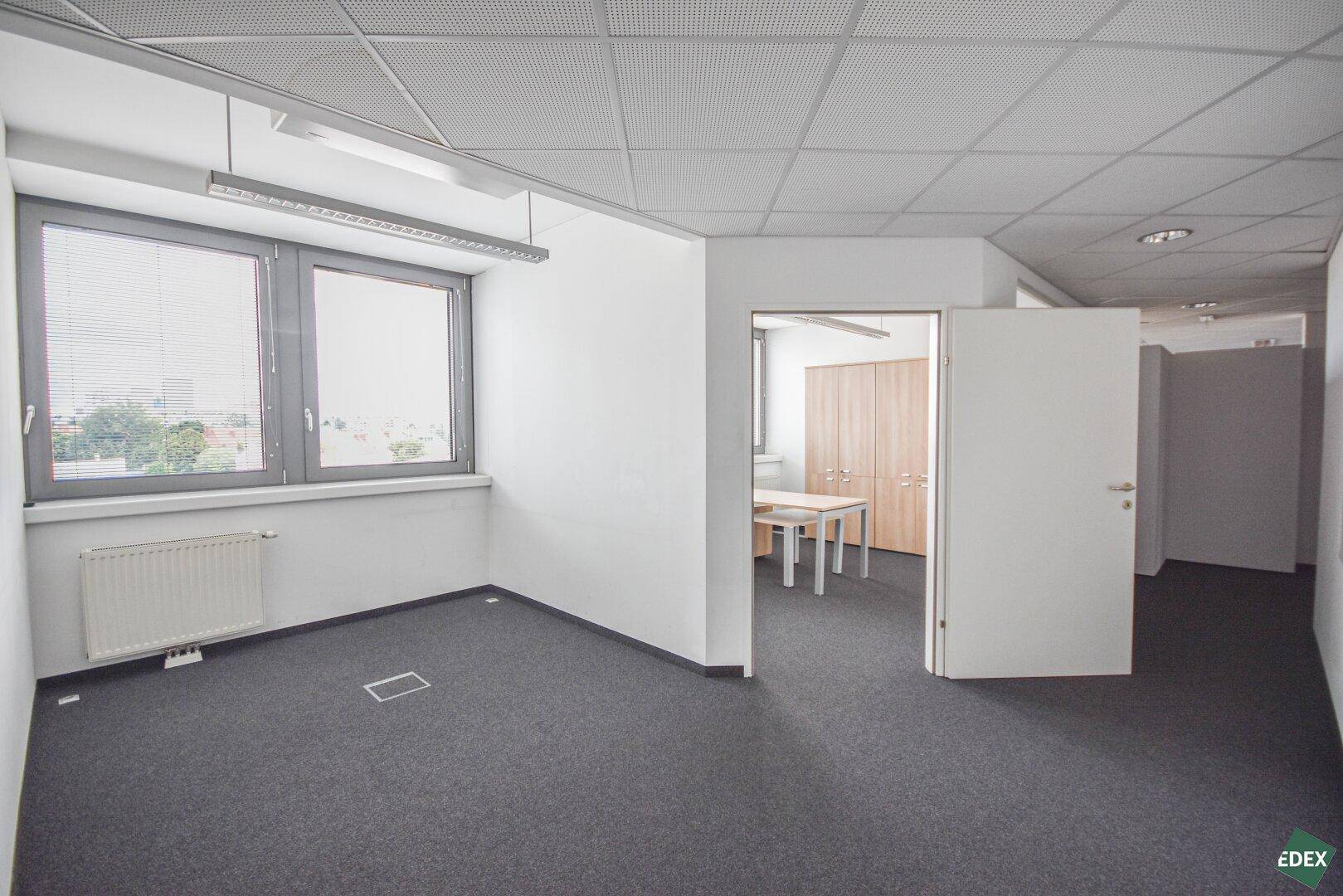 Heller Büroraum mit Teppichboden und Zugang zu einem weiteren Zimmer, ausgestattet mit Jalousien.