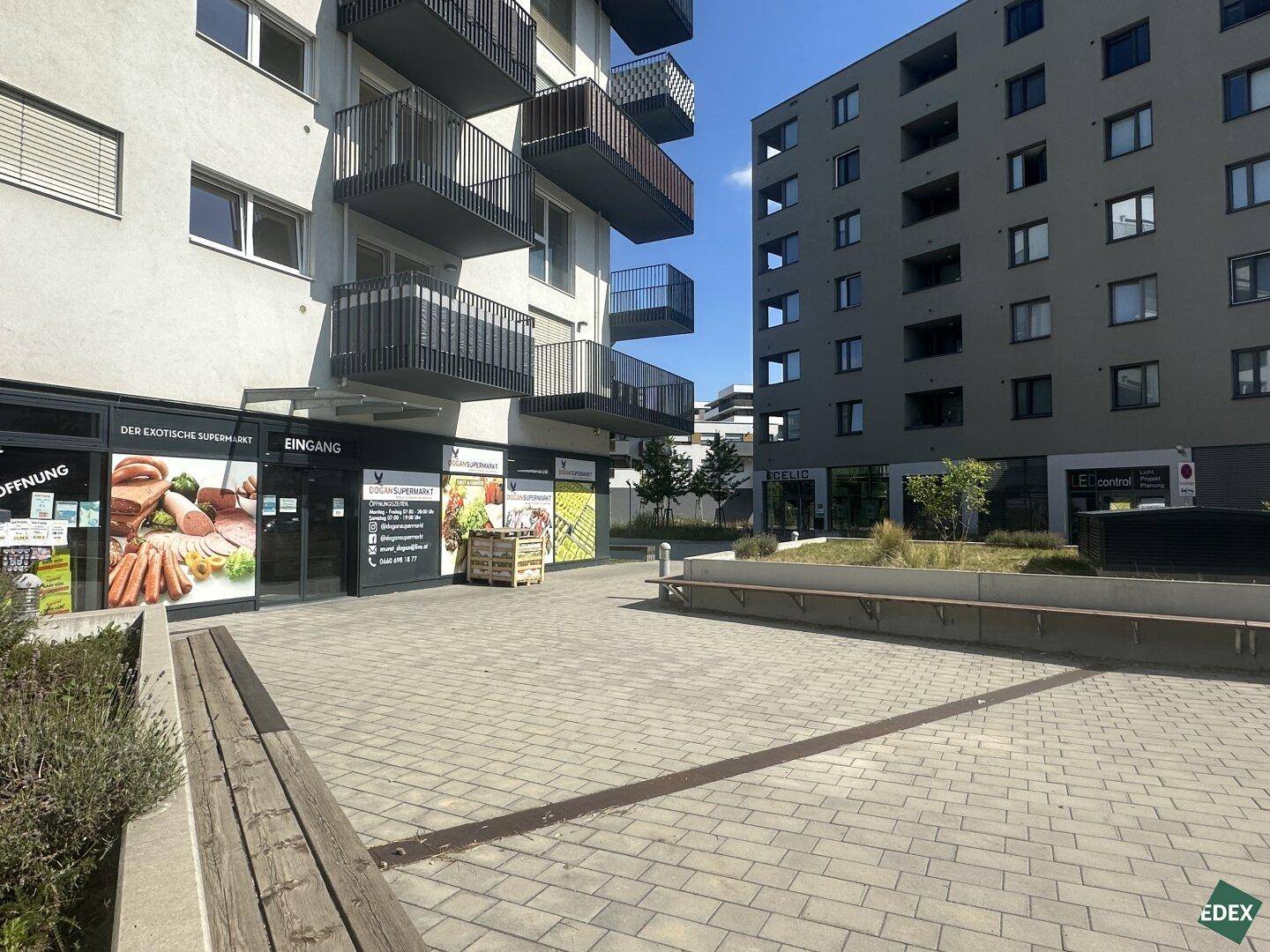 Blick auf die moderne Gebäudefassade mit Geschäften und einem gepflasterten Außenbereich mit Sitzbänken.