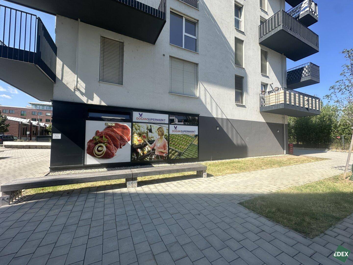 Detailansicht der unteren Gebäudefassade mit großen Schaufenstern eines Supermarktes und Sitzgelegenheiten.