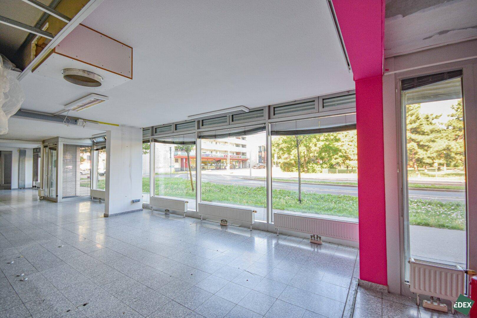Eingangsbereich des Gewerberaums mit Glasfront, rosa Wand und Blick auf die belebte Straße.