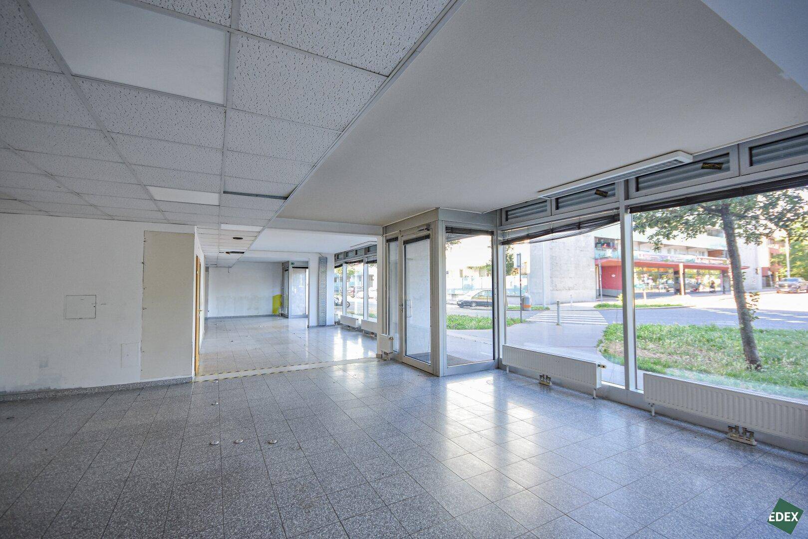 Großer, leerer Gewerberaum mit Fliesenboden, abgehängter Decke und großen Schaufenstern zur Straße.