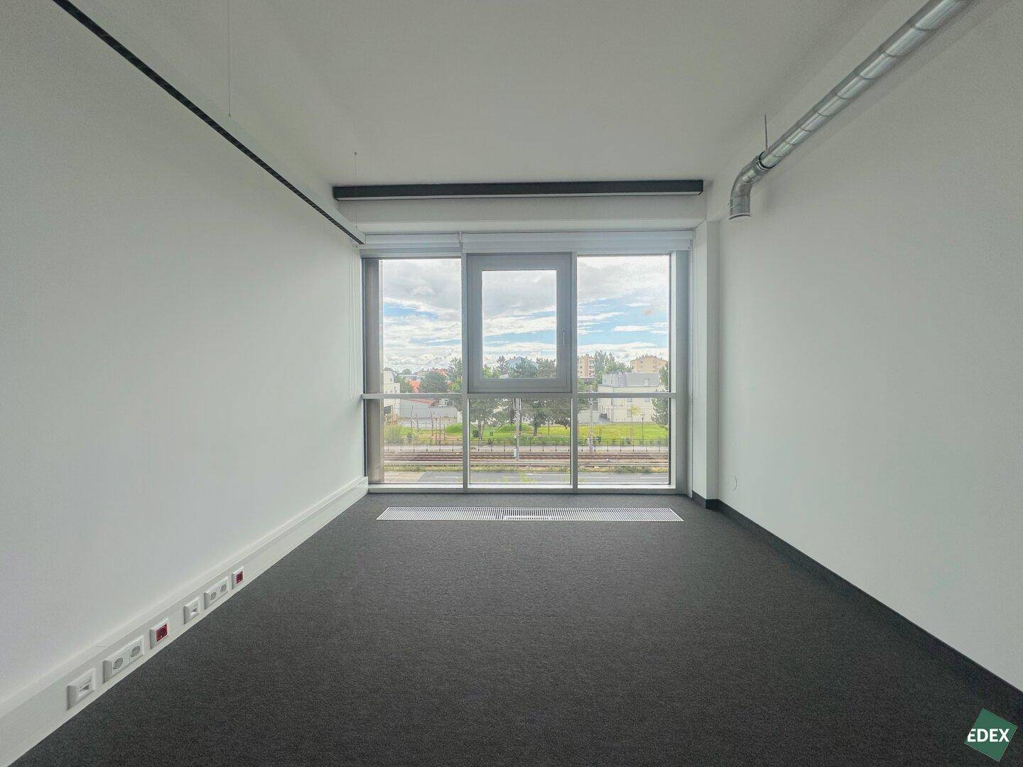 Leerer Büroraum mit dunklem Teppichboden und großem Fenster mit Blick auf Bahngleise und Bäume.