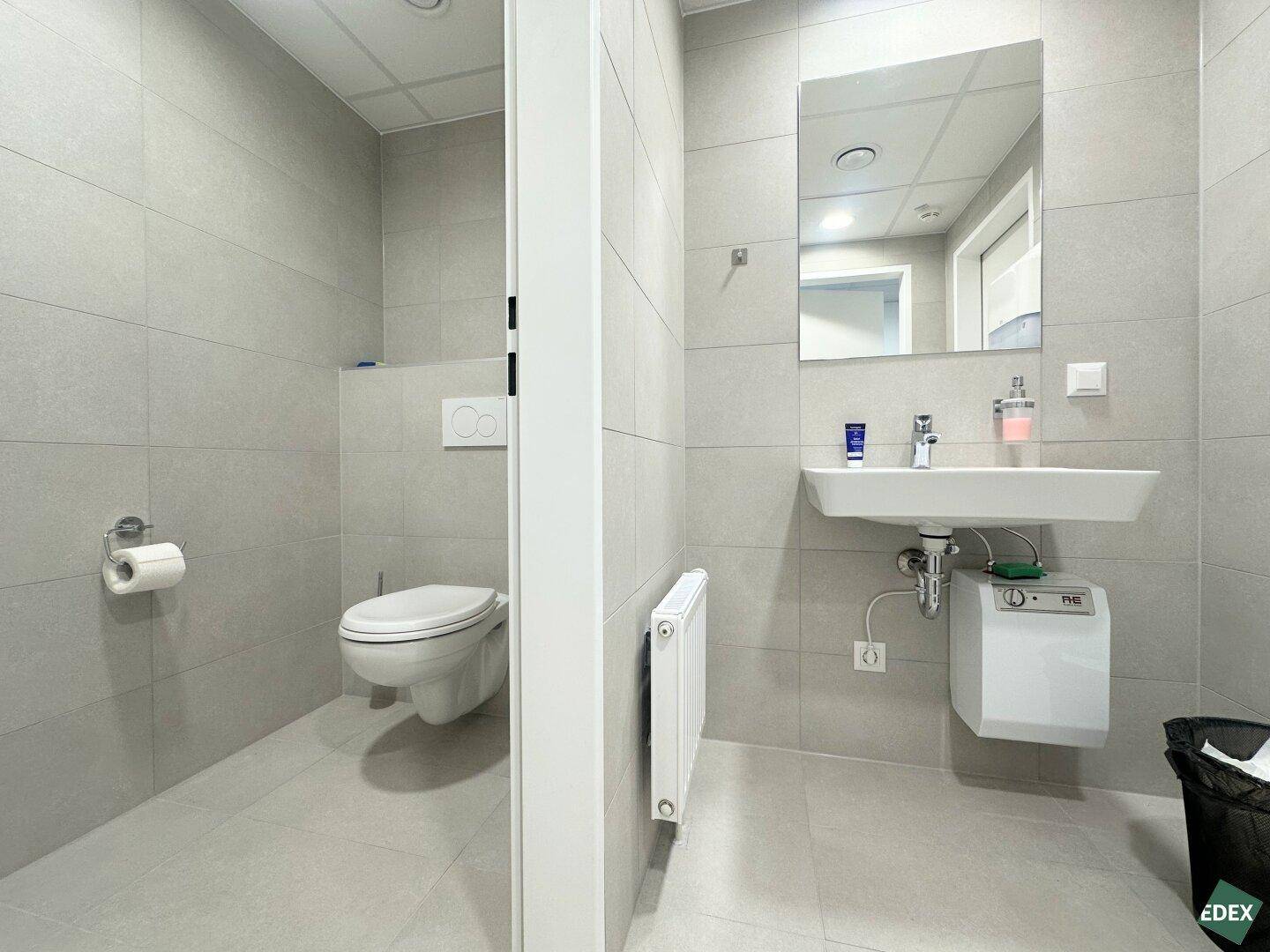 Modernes Badezimmer mit hellgrauen Fliesen, wandhängendem WC, Waschbecken und Heizkörper.