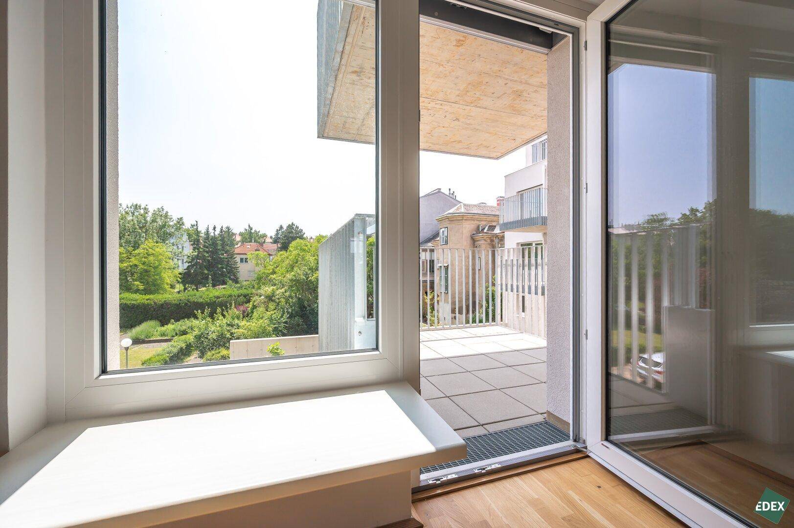 Blick aus dem Wohnzimmer durch große Fenster auf den Balkon und die grüne Umgebung.