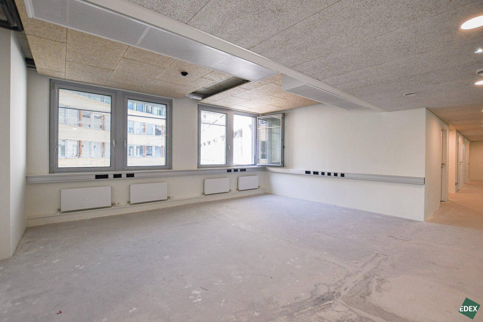 Unmöblierter Büroraum mit großen Fenstern, Akustikdecke und Wandanschlüssen für Technik.