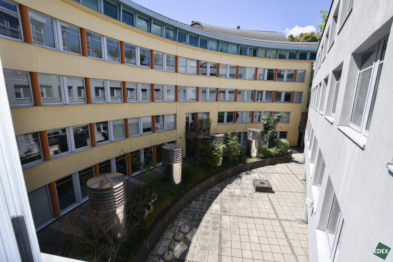 Innenhof eines modernen Bürogebäudes mit Bepflanzung und gepflasterter Fläche.