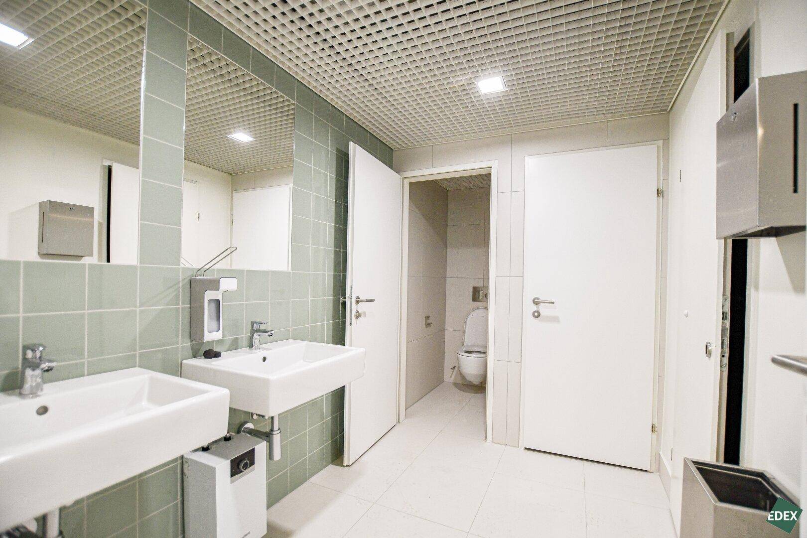 Modernes Badezimmer mit zwei Waschbecken, hellgrünen Fliesen und einem Gitterdeckenraster.