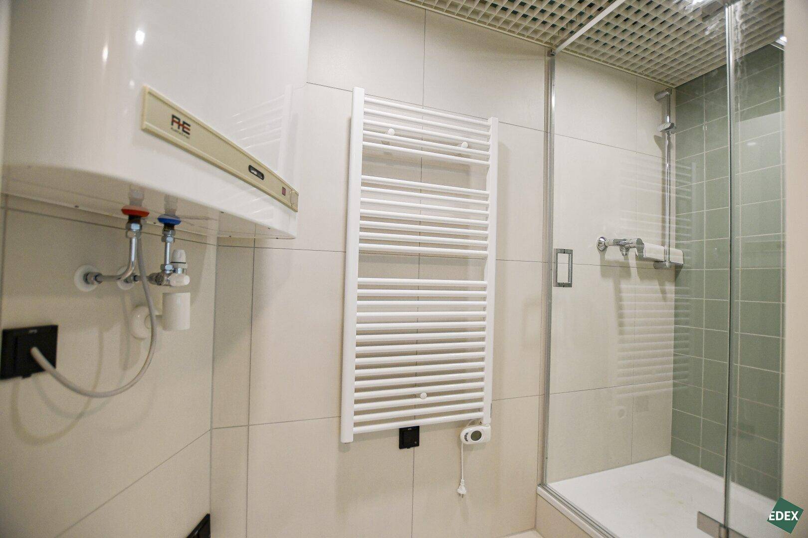 Badezimmer mit Duschkabine, Handtuchheizkörper und Warmwasserboiler an der Wand.