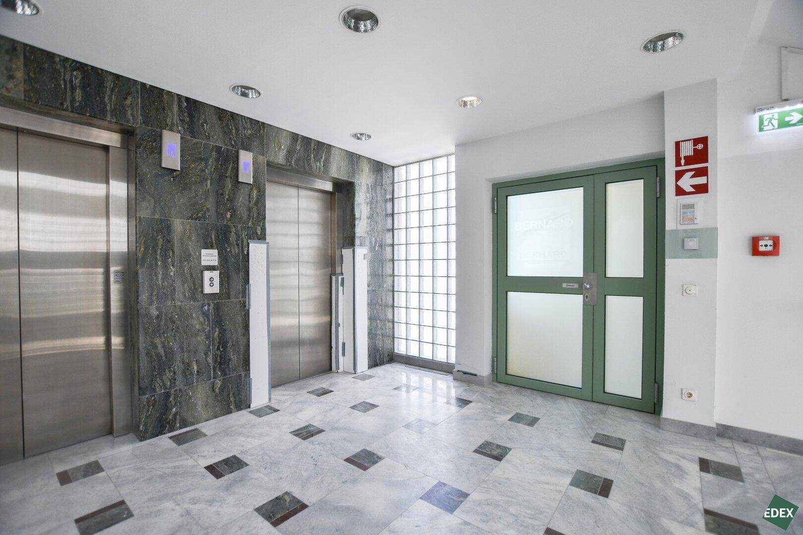 Moderner Eingangsbereich mit zwei Aufzügen, Marmorboden und einer Glastür zum Treppenhaus.