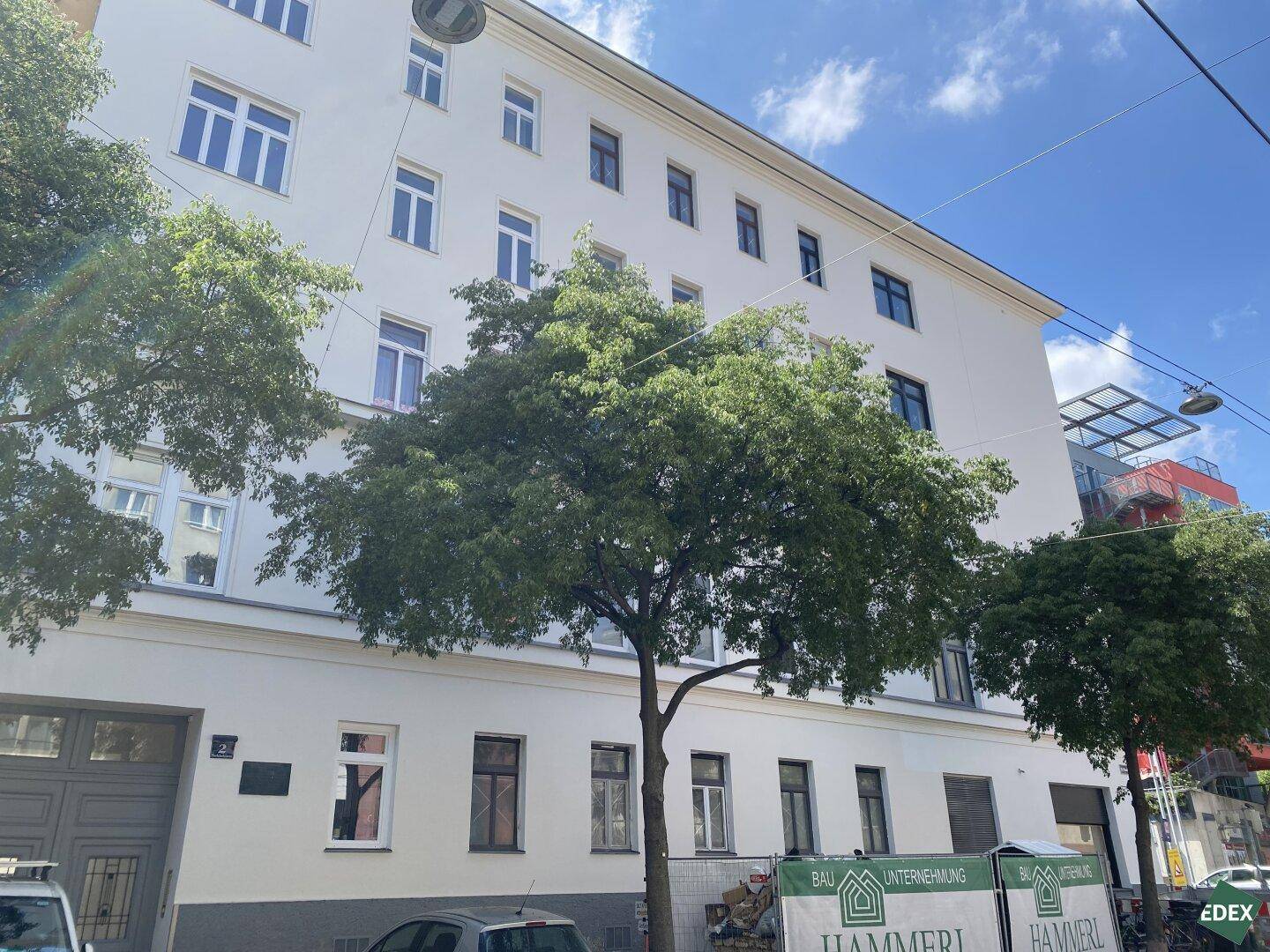 Ansicht der Gebäudefassade von der Straße mit Bäumen und Bauplakaten im Vordergrund.