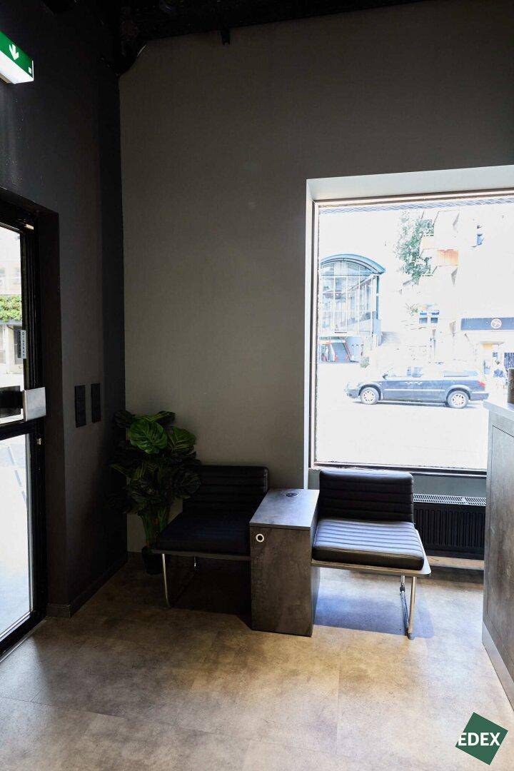 Gemütlicher Wartebereich mit zwei Sitzgelegenheiten und einem Beistelltisch direkt am großen Fenster zur Straße.