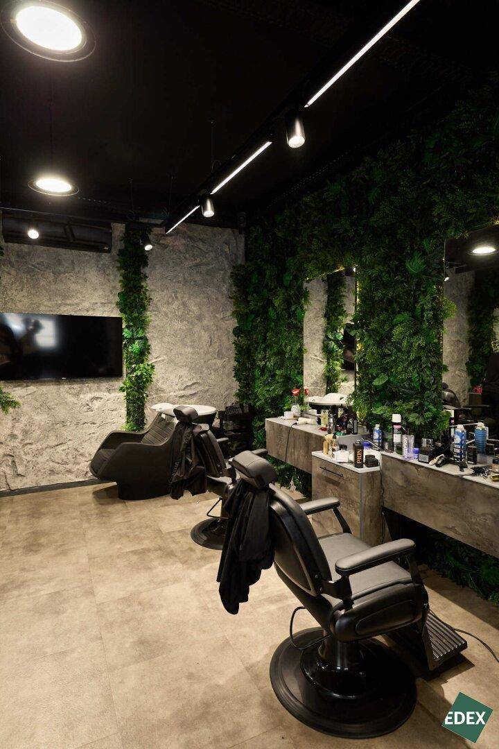 Moderner Friseurbereich mit Stylingstühlen, Waschbecken, Fernseher und Wänden, die mit Kunstpflanzen und Steinoptik gestaltet sind.