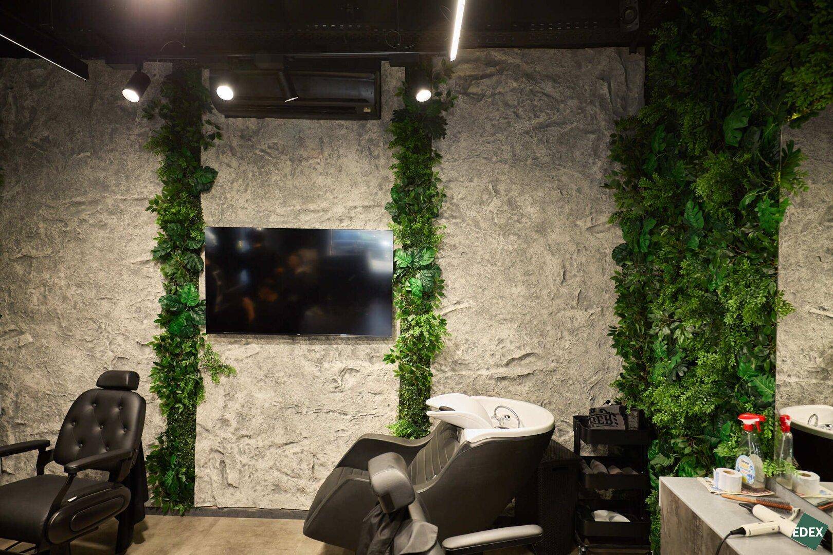 Detailansicht des Friseurbereichs mit strukturierter Wand, vertikalen Kunstpflanzen, Fernseher und Haarwaschbecken.