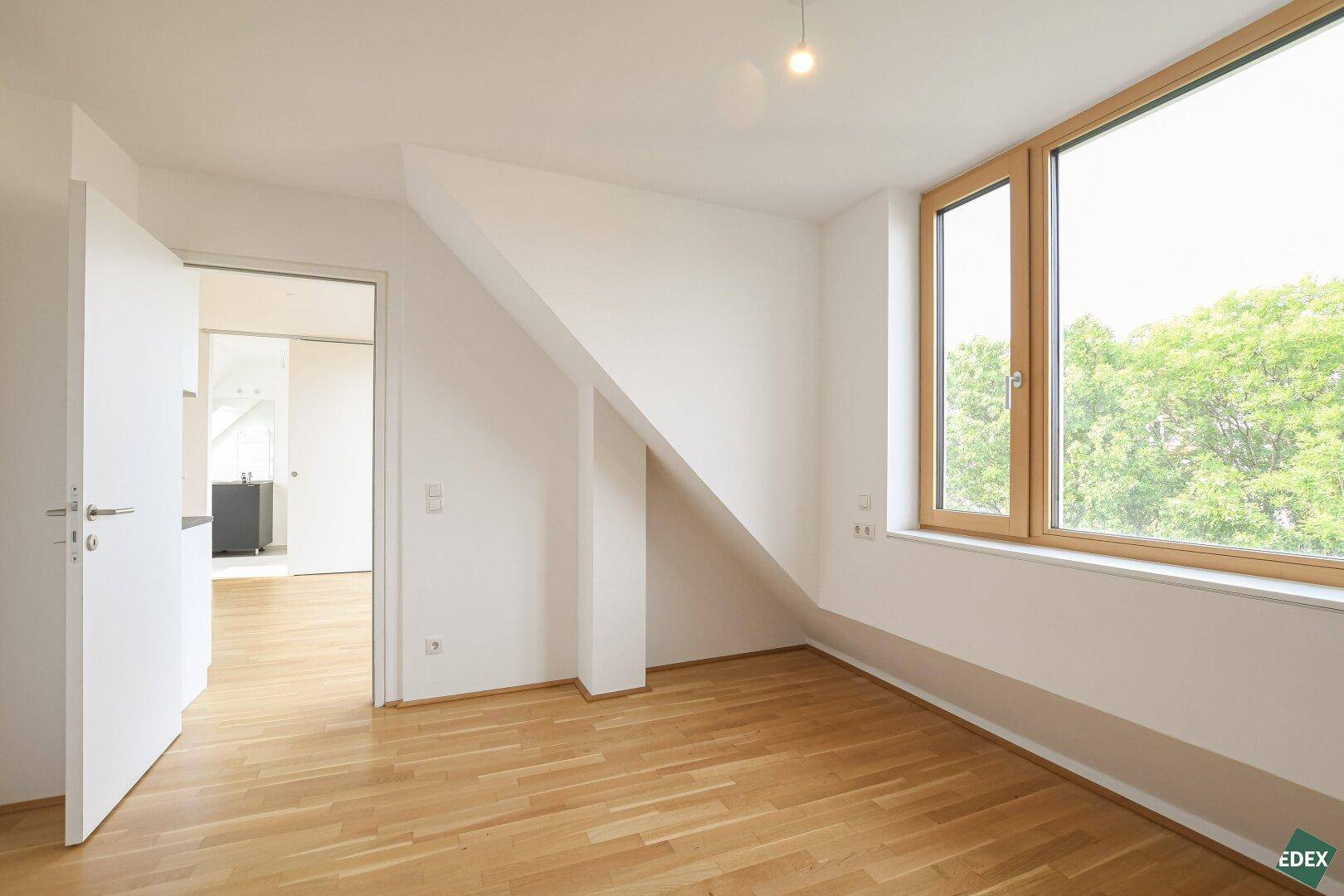 Gemütliches Zimmer mit Dachschräge, Parkettboden und großen Fenstern mit Blick ins Grüne.