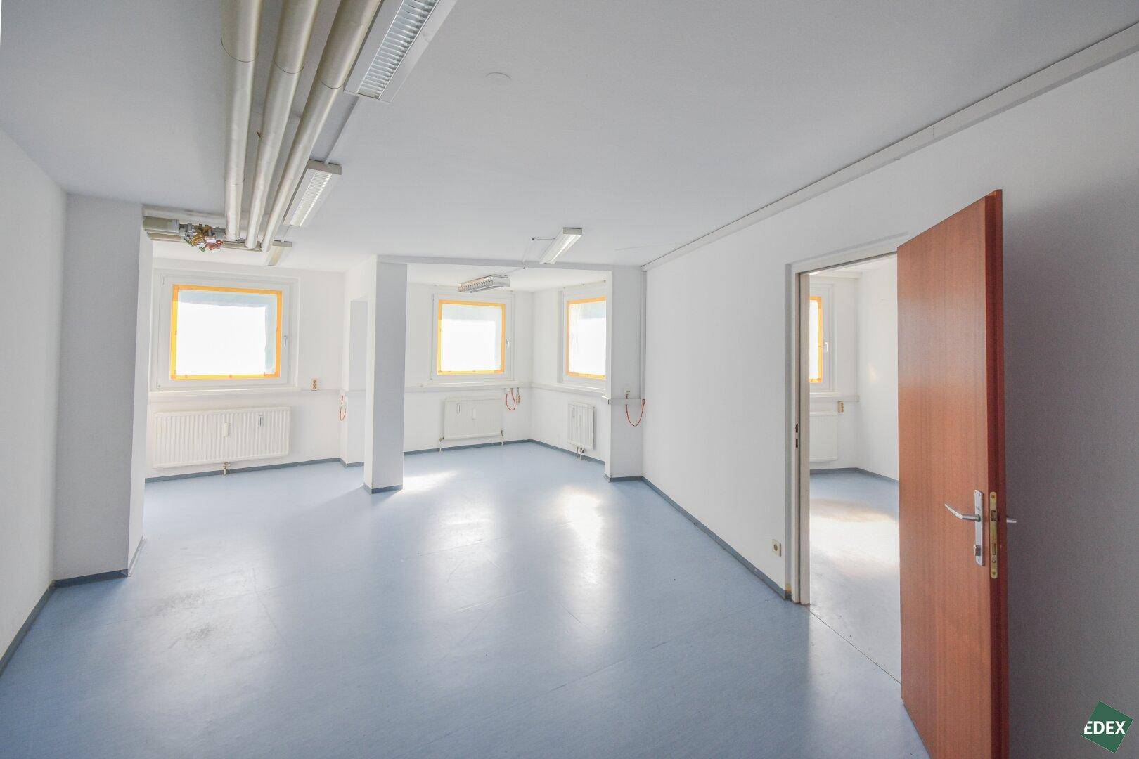 Geräumiger Bürobereich mit blaugrauem Boden, weißen Wänden und sichtbaren Heizungsrohren an der Decke.