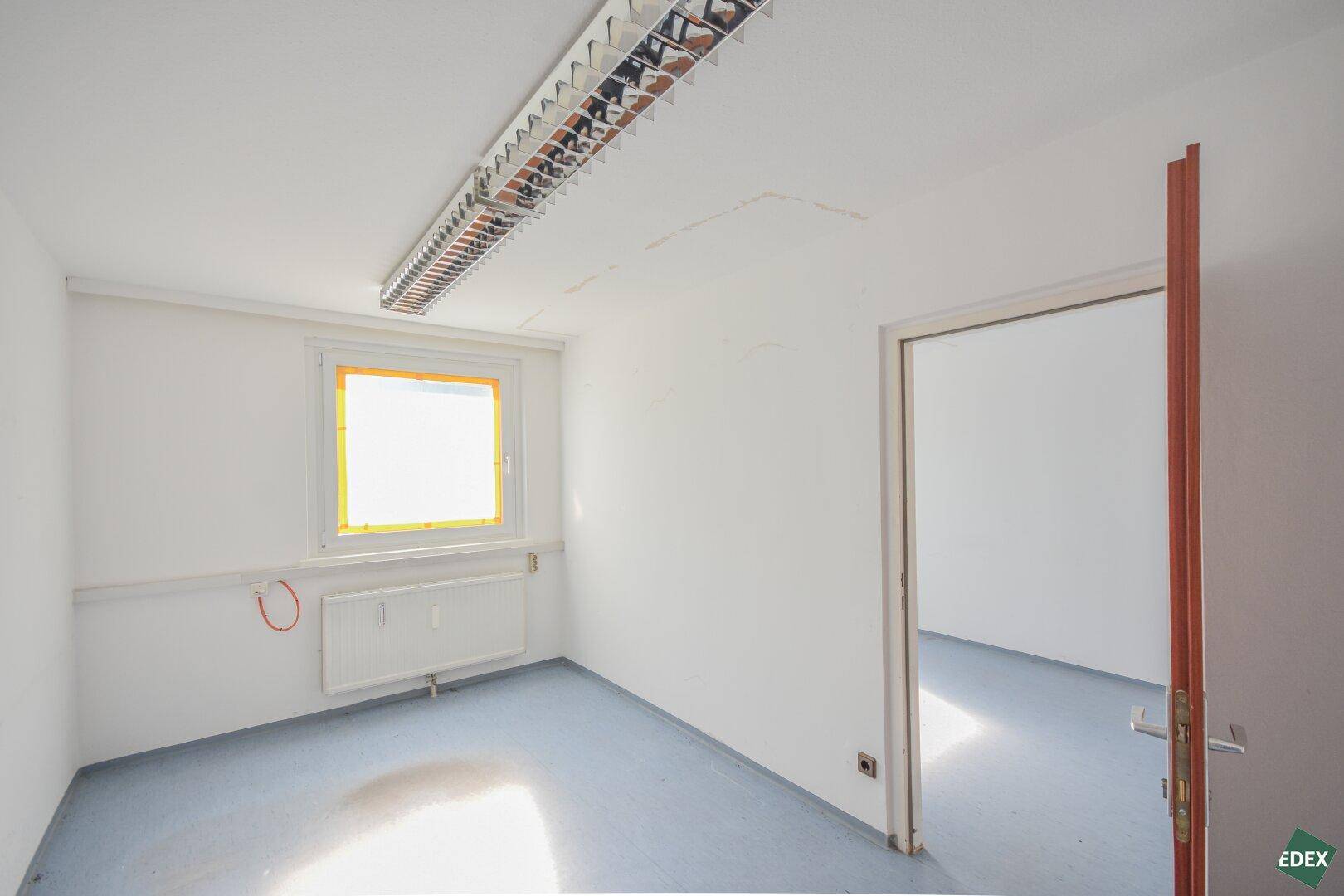 Einzelner Büroraum mit blaugrauem Boden, weiß gestrichenen Wänden und einem Fenster mit Heizkörper.