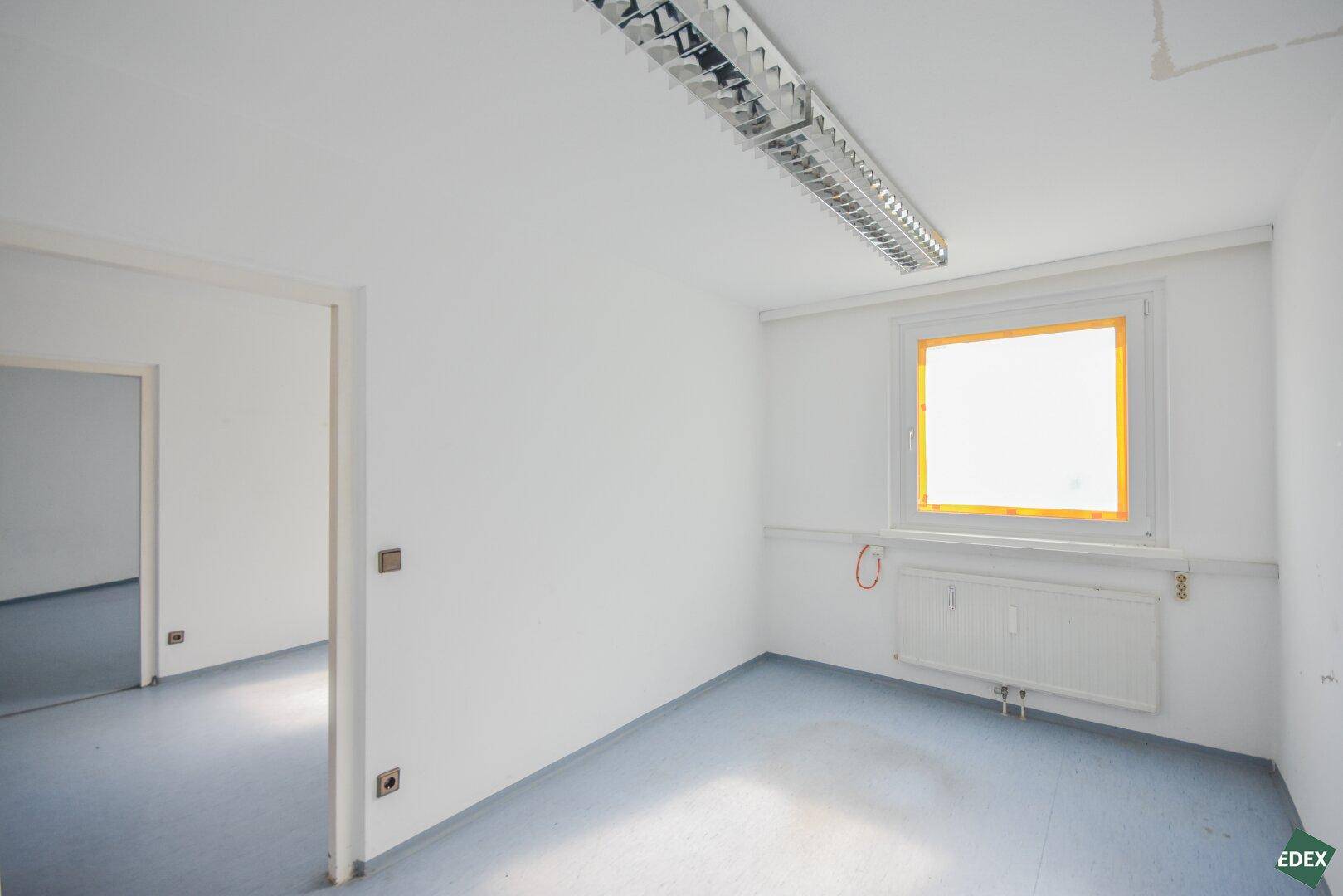 Büroraum mit blaugrauem Boden, weißen Wänden und einem Fenster, das den Raum erhellt.
