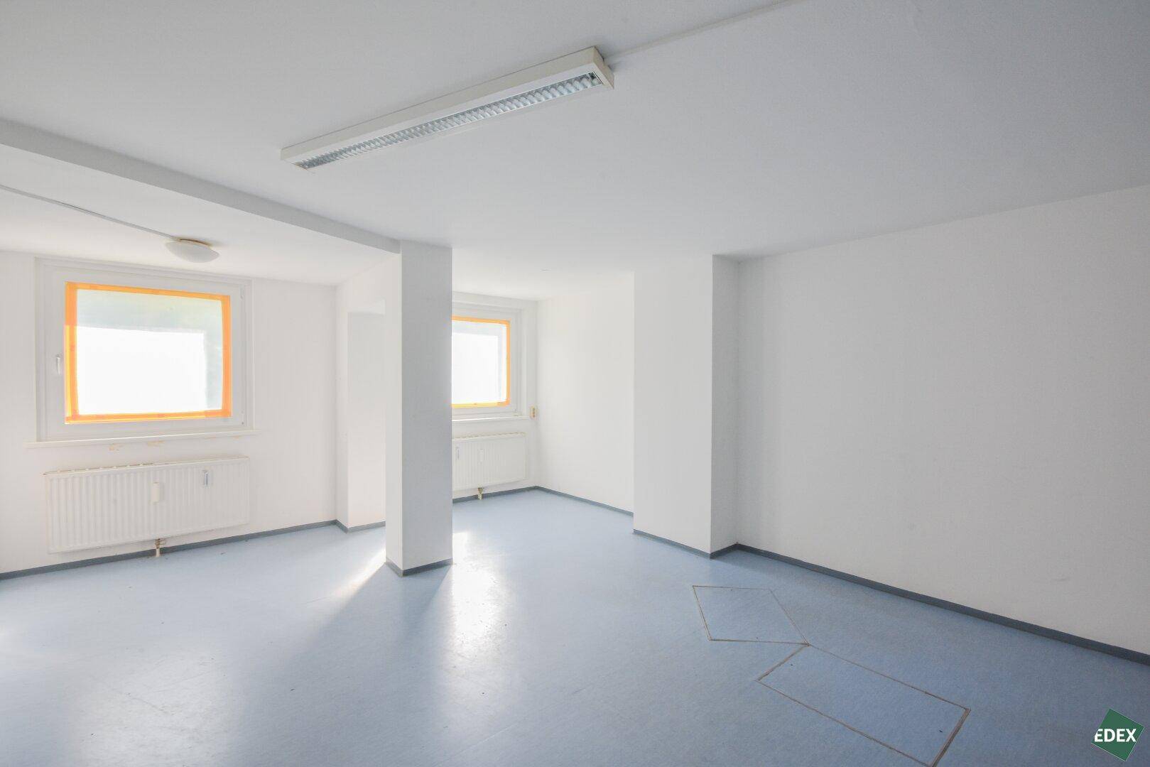 Heller Büroraum mit blaugrauem Boden, weißen Wänden und zwei Fenstern, die den Raum mit Tageslicht füllen.