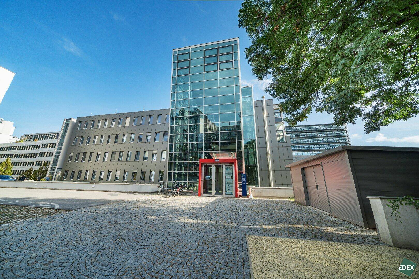 Eingangsbereich eines modernen Bürogebäudes mit Glasfassade und gepflastertem Vorplatz.