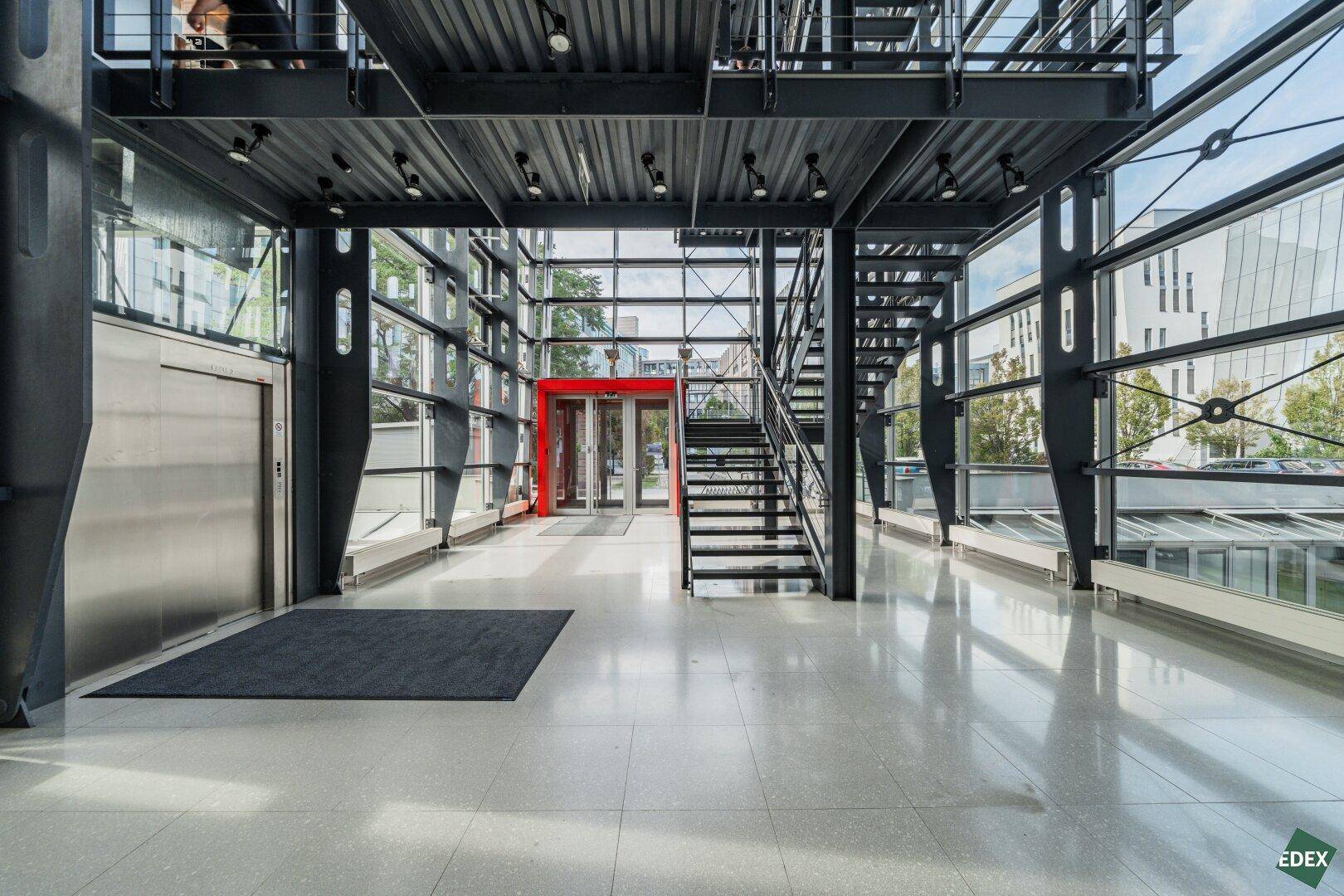 Heller Eingangsbereich mit Aufzug, moderner Treppe und großen Glasflächen.