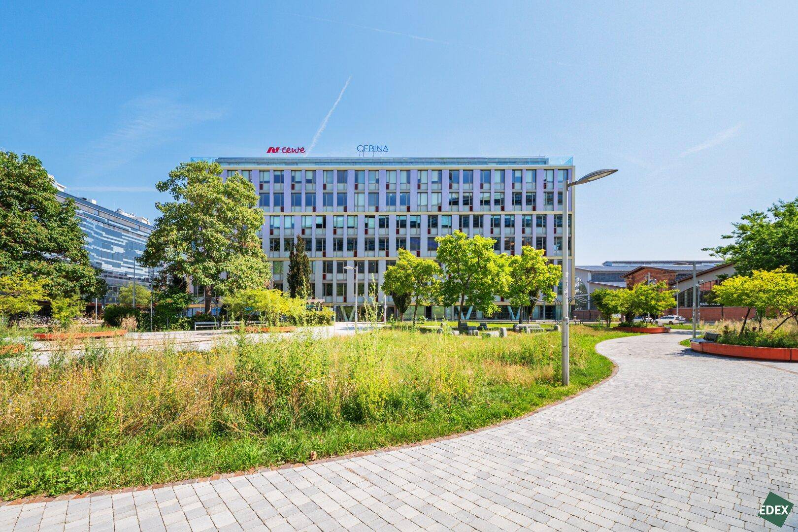 Modernes Bürogebäude mit Glasfassade, umgeben von Grünflächen und Bäumen unter blauem Himmel.