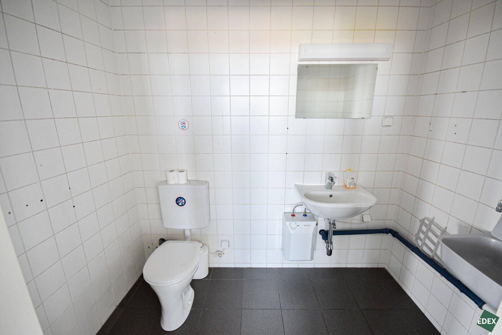 Einfache Toilettenanlage mit weißen Wandfliesen, WC, Waschbecken und Spiegel.