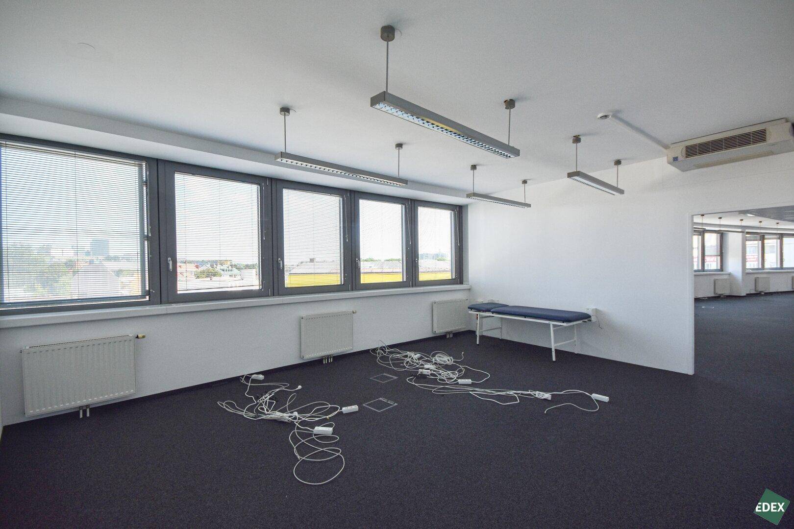 Heller Büroraum mit Teppichboden, großen Fenstern und einer Untersuchungsliege im Vordergrund.