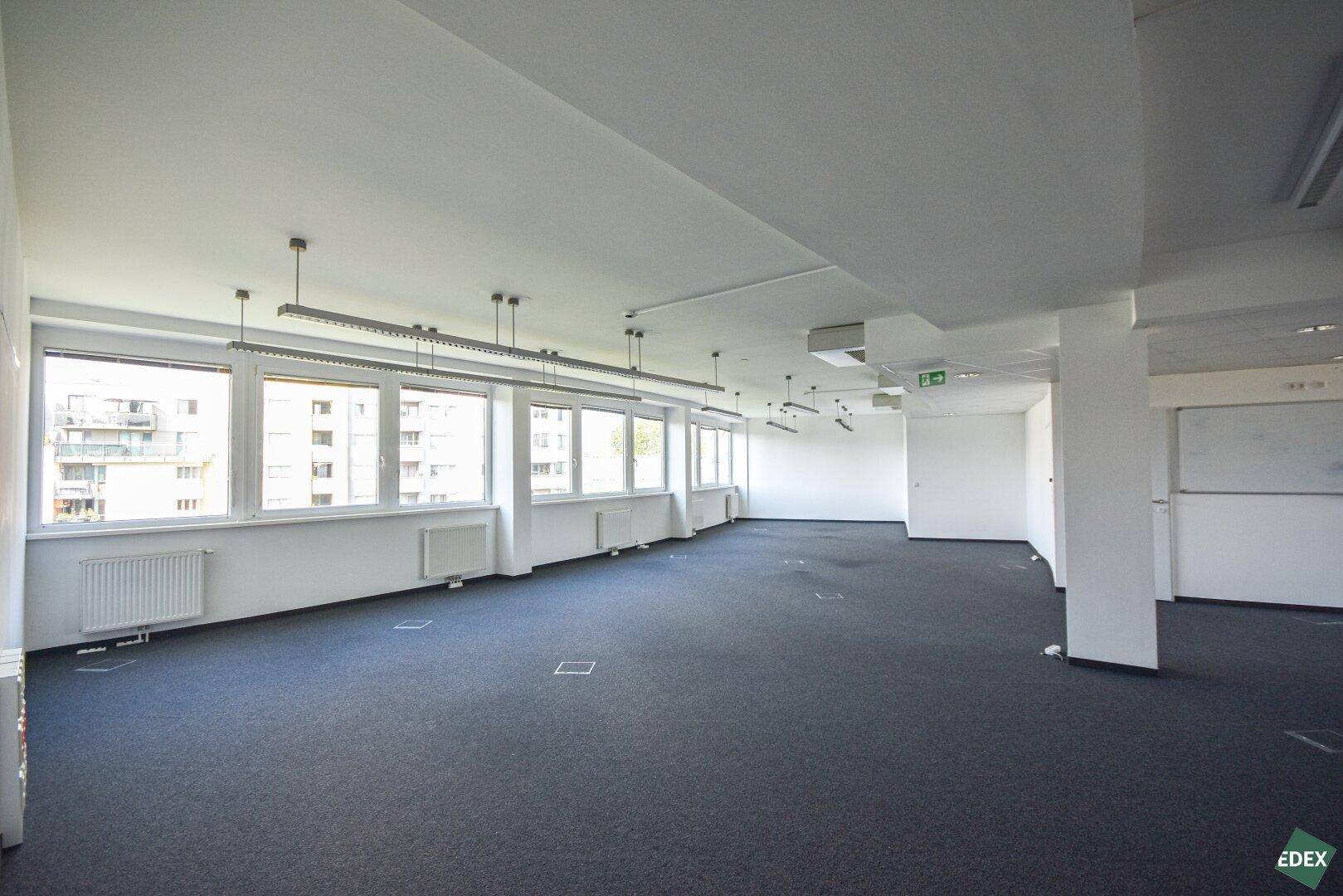 Weitläufiger Bürobereich mit Teppichboden, vielen Fenstern und einer Säule im Hintergrund.