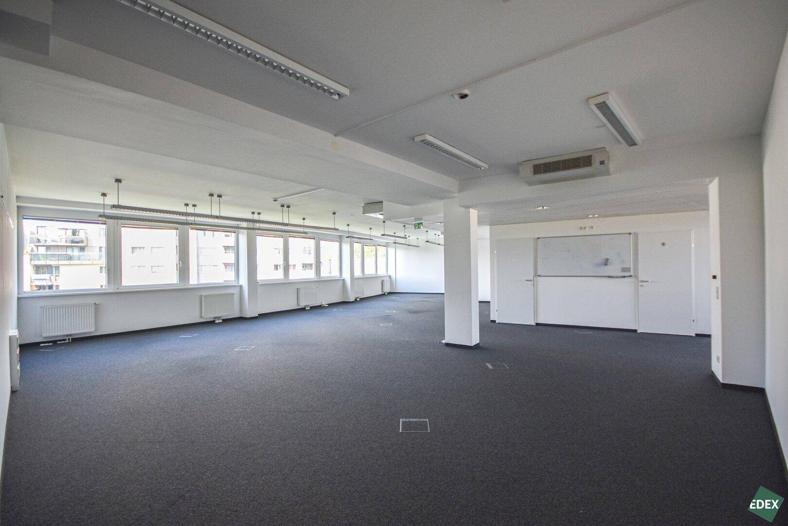 Großer, unmöblierter Bürobereich mit Teppichboden, Säulen und einer Whiteboard-Wand.