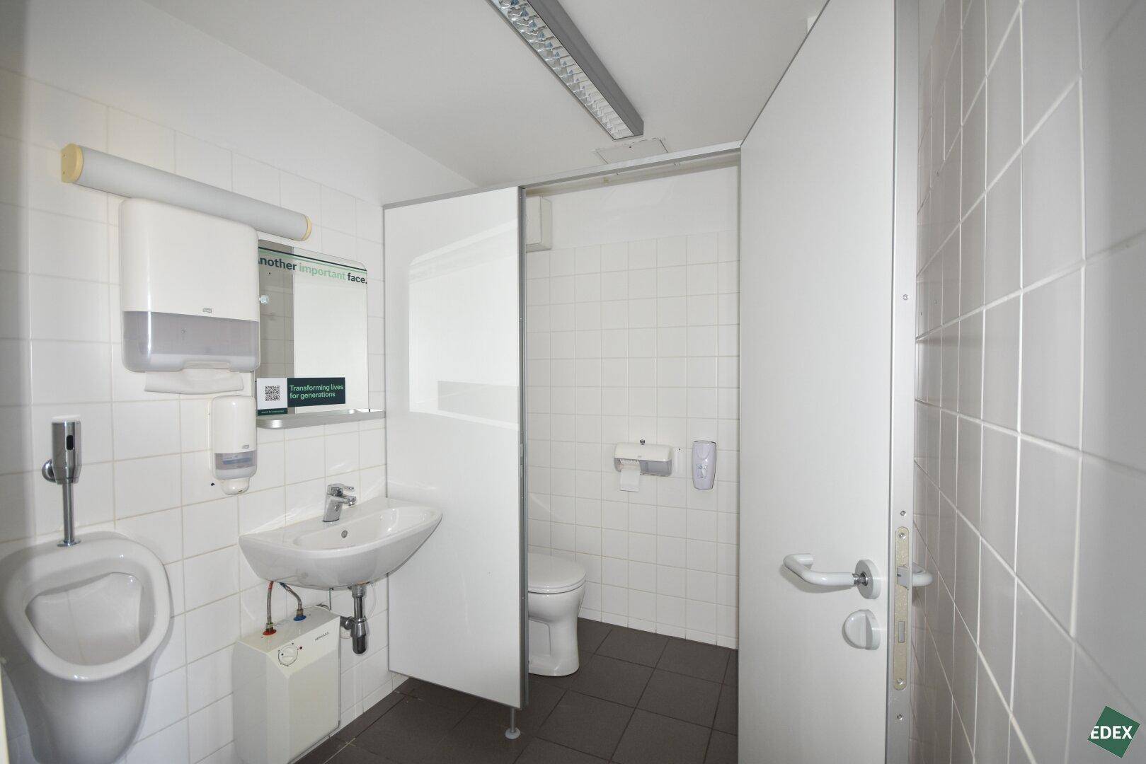 Helle Toilettenanlage mit weißen Fliesen, Urinal, Waschbecken und einer Toilettenkabine.