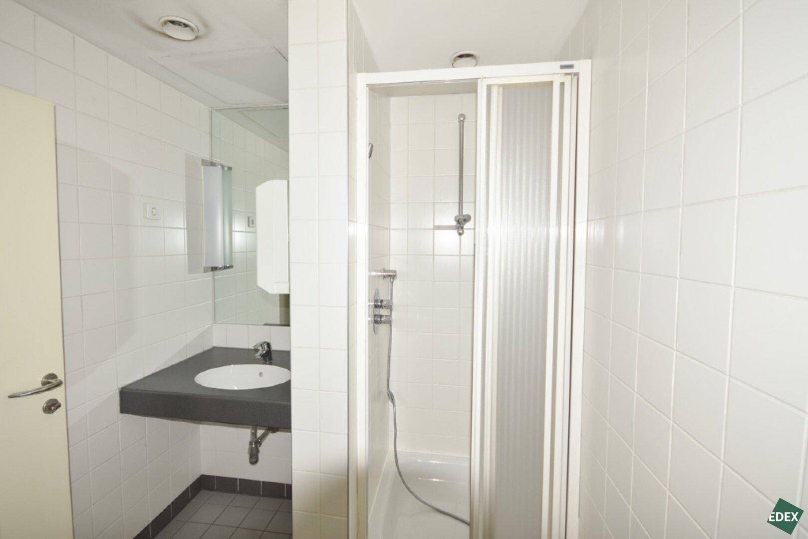 Helles Badezimmer mit weißen Fliesen, einer Duschkabine und einem modernen Waschbecken mit dunkler Arbeitsplatte.