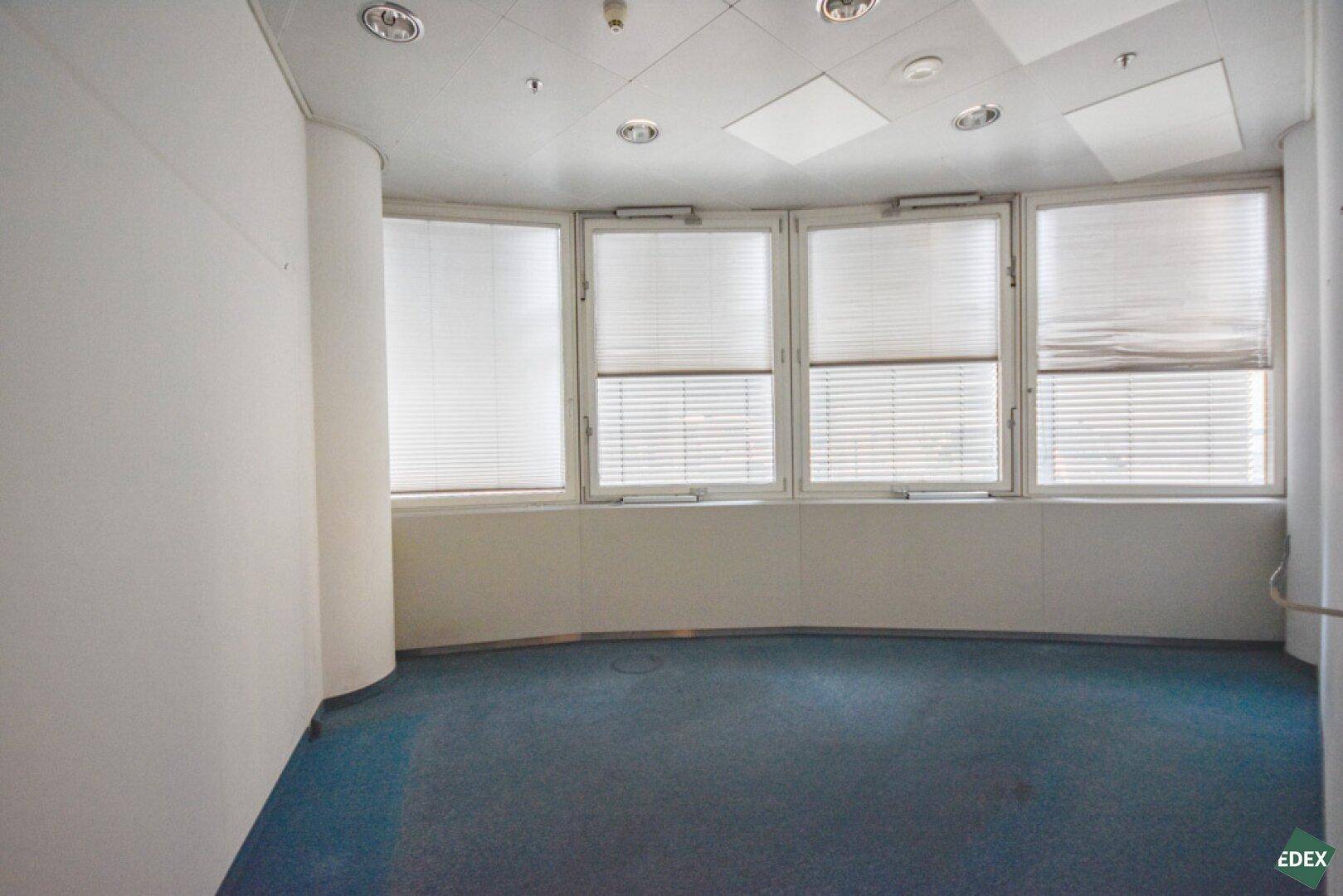 Kleinerer Büroraum mit blauem Teppichboden, Fensterjalousien und einer geschwungenen Wand, ideal für Besprechungen.