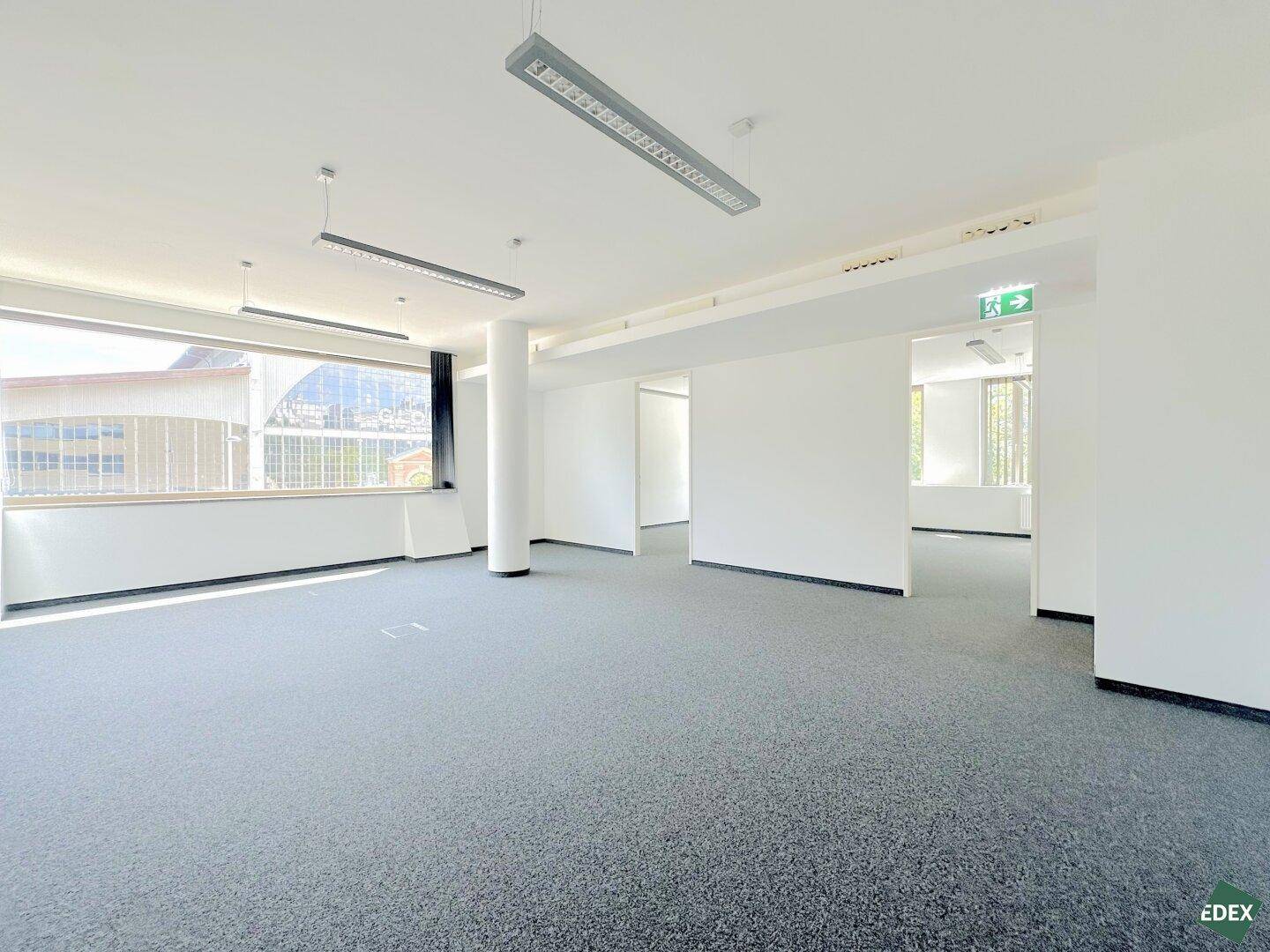 Offener Bürobereich mit Teppichboden, großen Fenstern und Blick in angrenzende Räume.