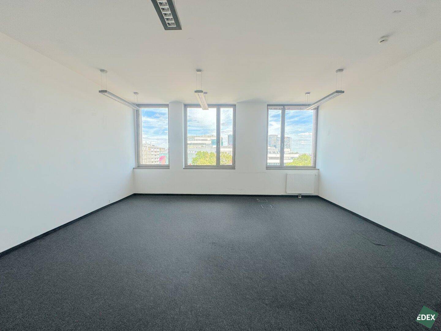Heller Büroraum mit Teppichboden, Heizkörper unter den Fenstern und Blick auf die Stadt.