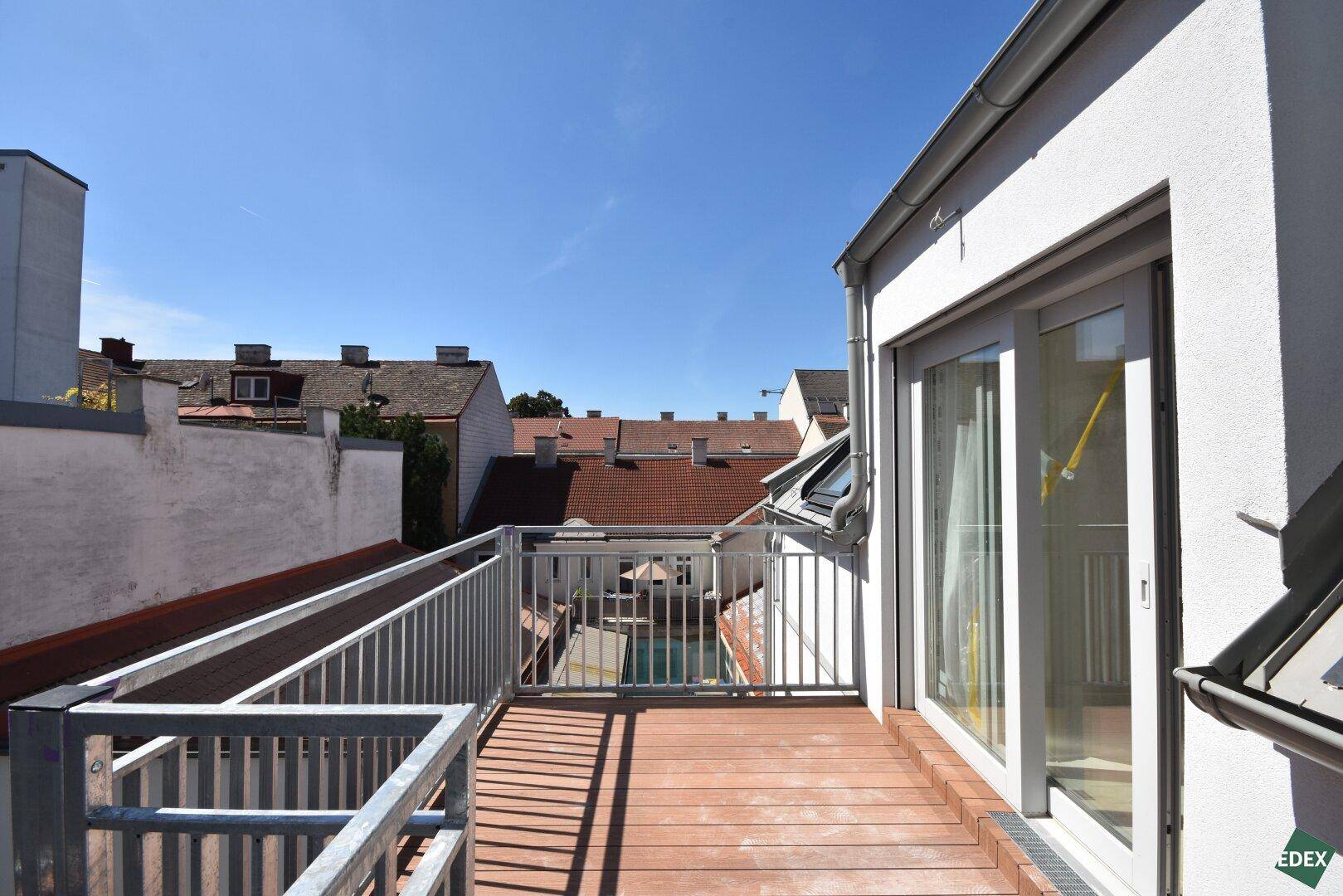 Sonniger Balkon mit Holzdielen und Metallgeländer, bietet einen Blick über die umliegenden Dächer.