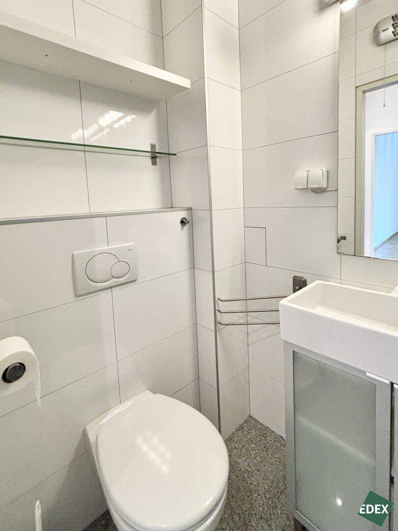 Helles WC mit wandhängender Toilette, Waschbecken und weißen Fliesen.