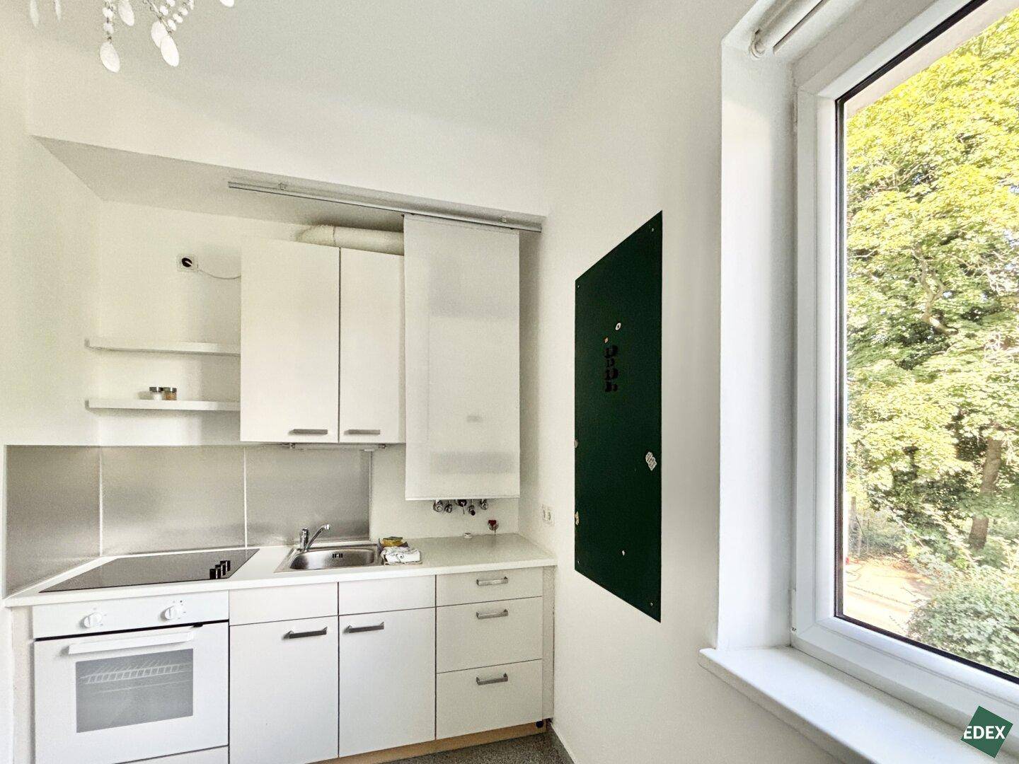 Kleine Küchenzeile mit weißen Schränken, Spüle und Herd, neben einem Fenster mit Grünblick.