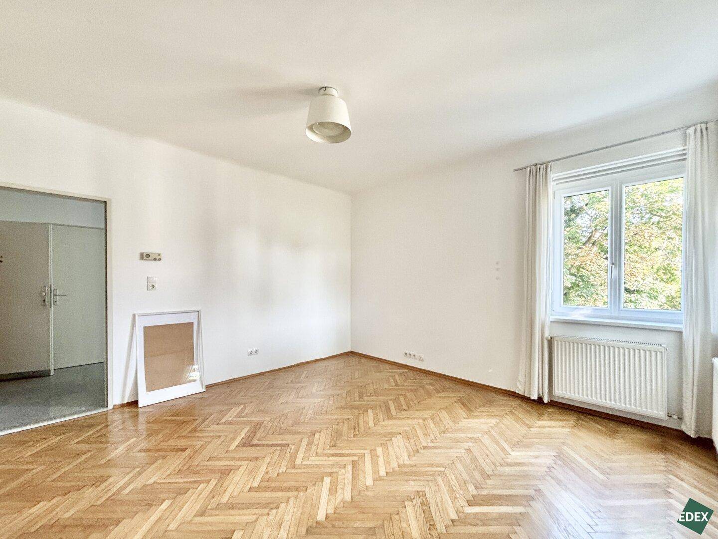 Geräumiges Zimmer mit Fischgrätparkett, weiß gestrichenen Wänden und einem Fenster.