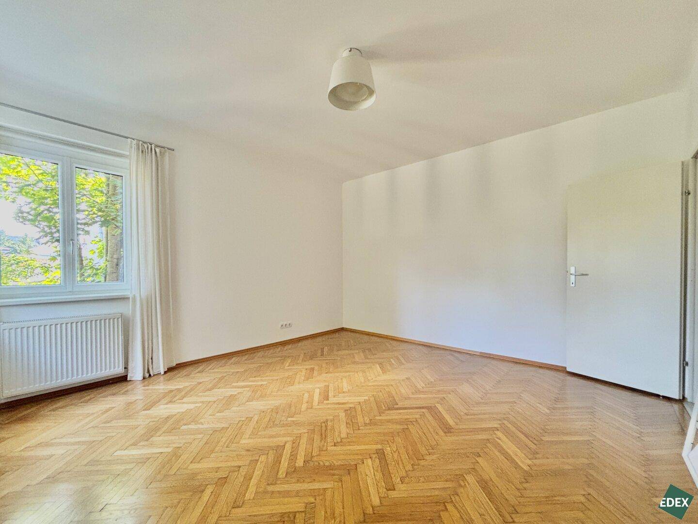 Helles Zimmer mit Fischgrätparkett, weißer Decke und einem Fenster mit Blick ins Grüne.