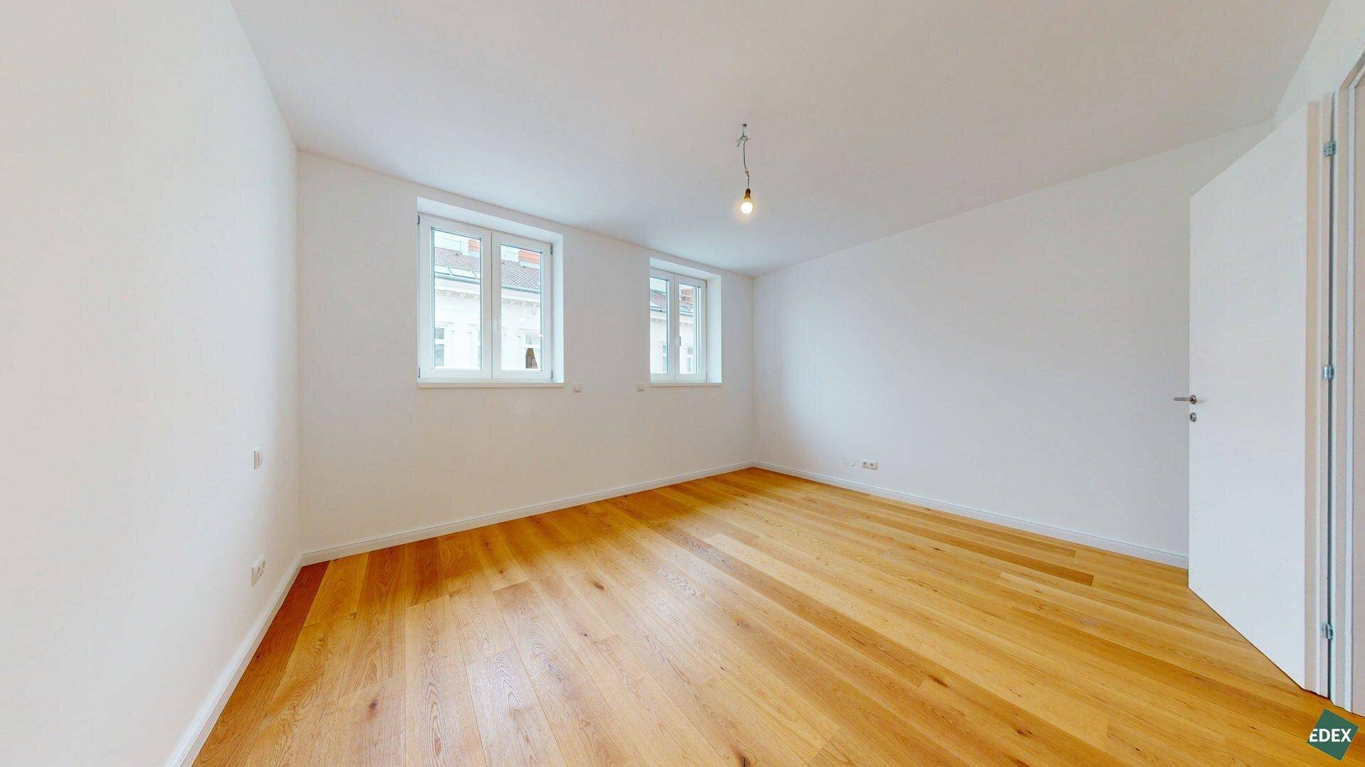 Helles Zimmer mit Holzboden und zwei Fenstern, die viel Tageslicht hereinlassen.