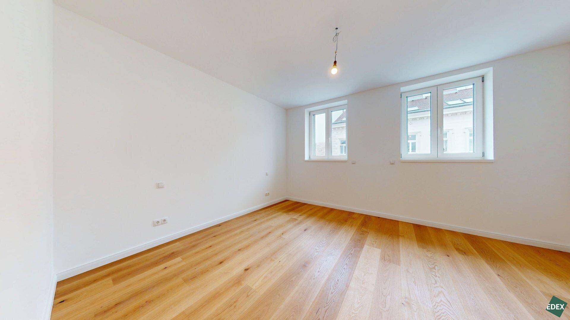 Geräumiges Zimmer mit Holzboden und zwei Fenstern, das eine angenehme Wohnatmosphäre bietet.