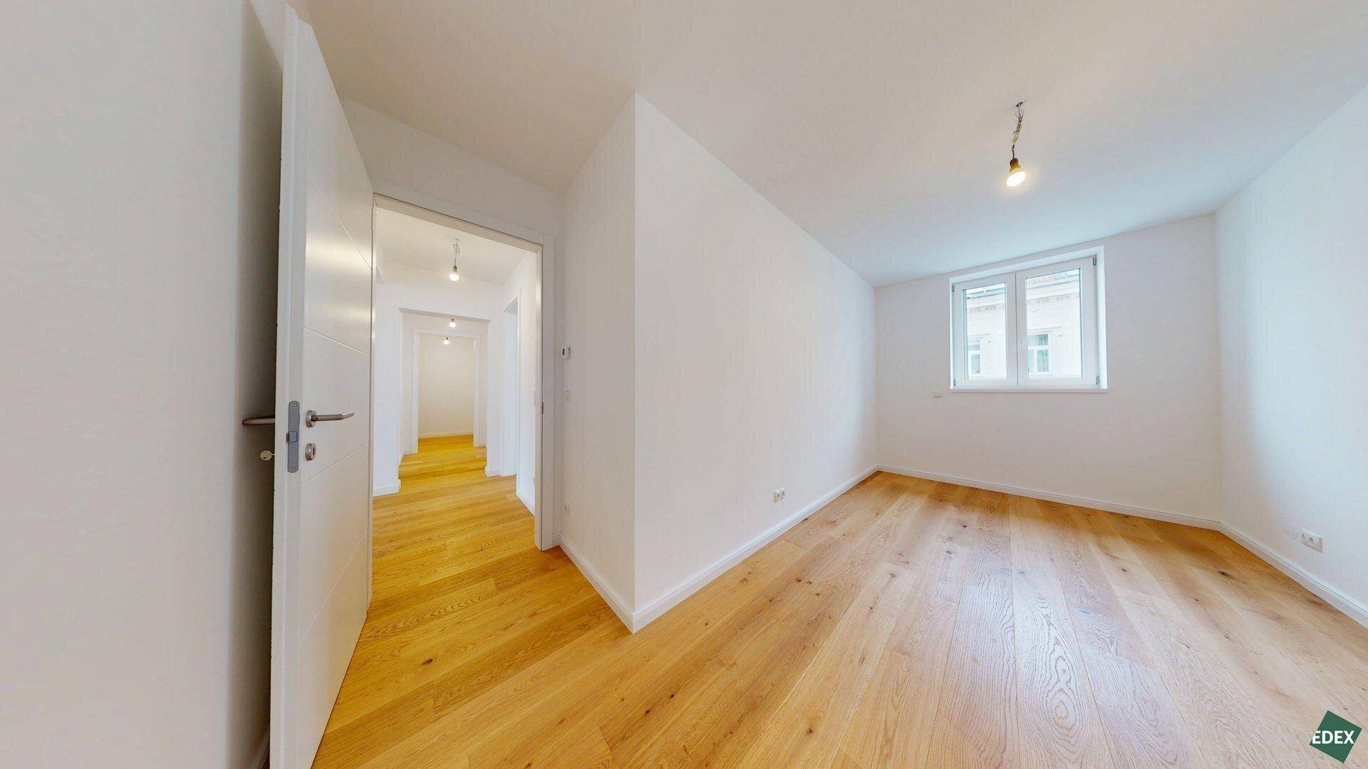 Helles Zimmer mit Holzboden und Fenster, das einen Zugang zu einem Flur bietet.