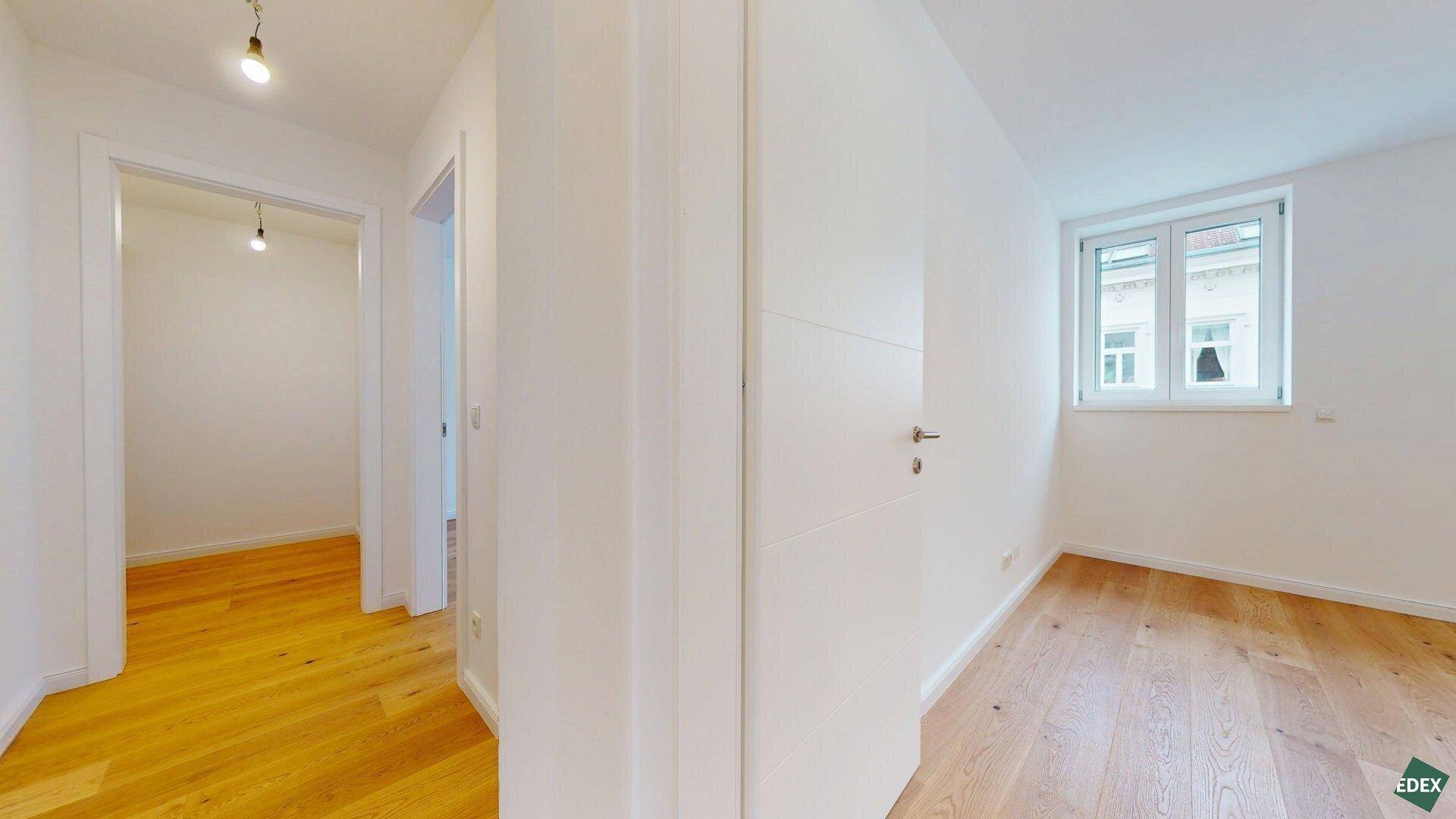Heller Flur führt zu einem Zimmer mit Holzboden und Fenster, das viel Tageslicht bietet.