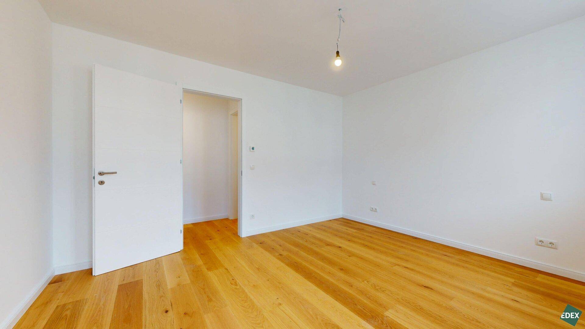 Leeres Zimmer mit hellem Holzparkettboden und einer Tür, die zu einem weiteren Bereich führt.
