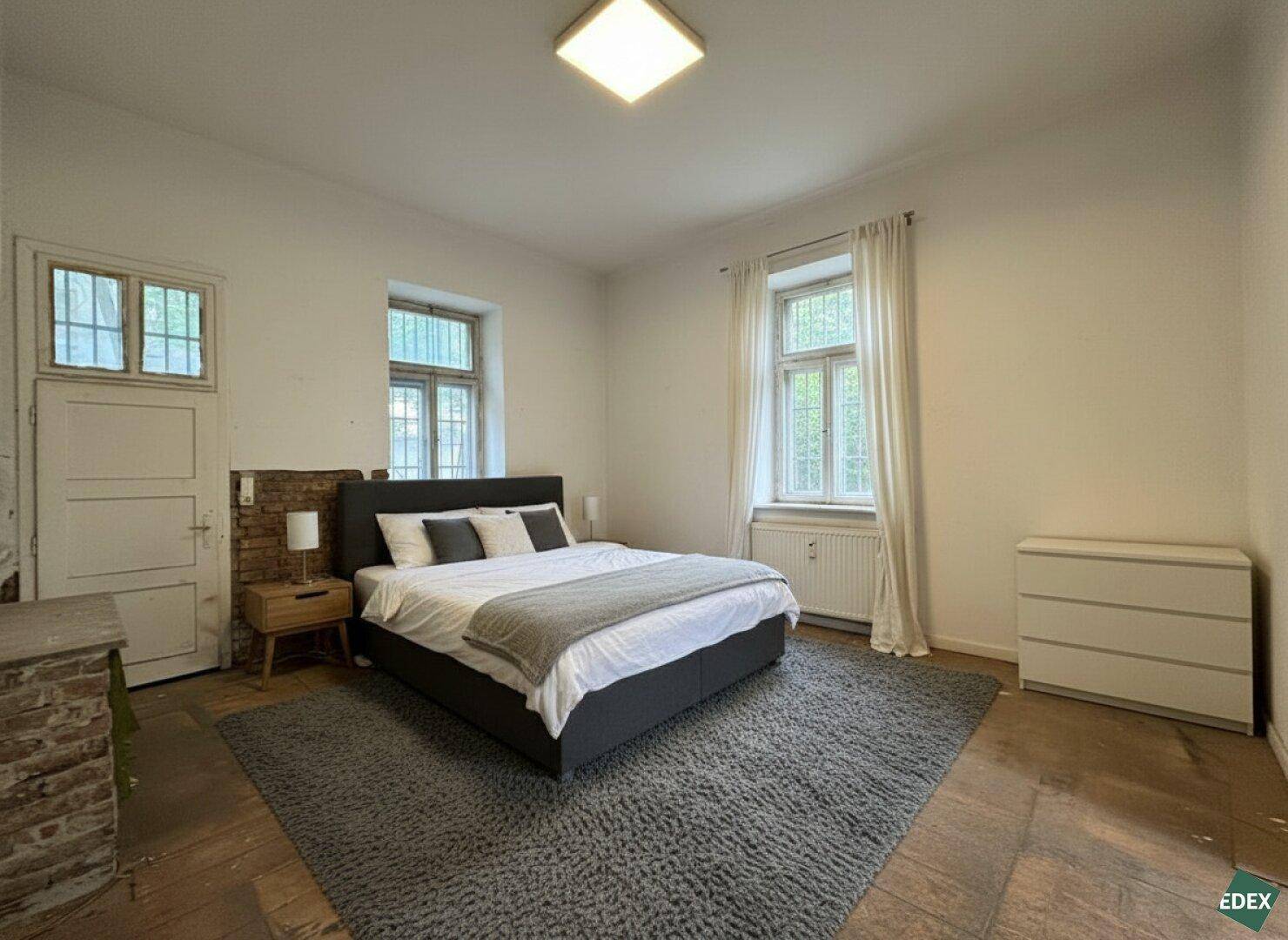 Schlafzimmer mit Doppelbett, Teppich und zwei Fenstern, die viel Licht hereinlassen.