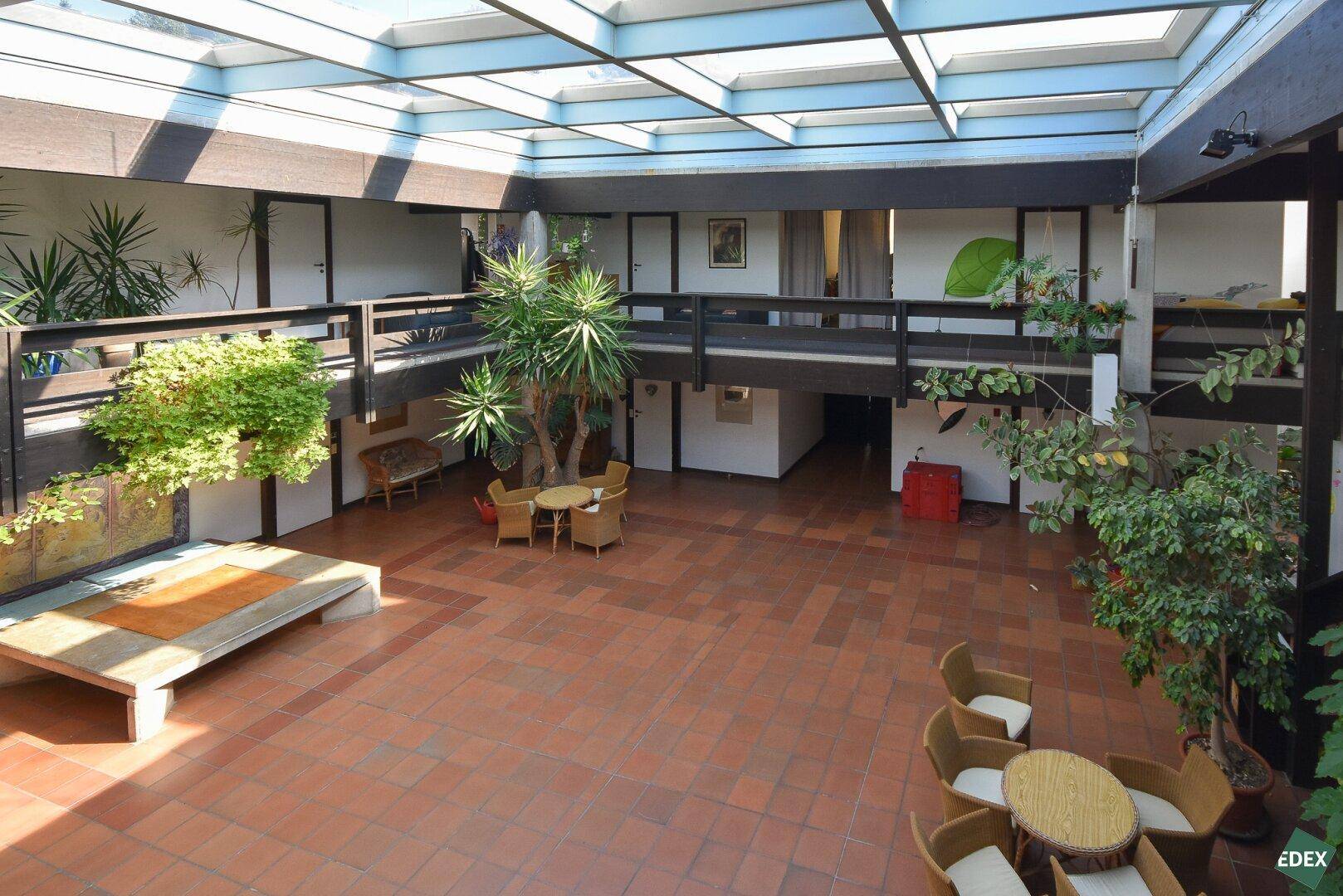 Heller Innenhof mit Glasdach, Terrakottafliesen und Pflanzen, umgeben von Galerien.