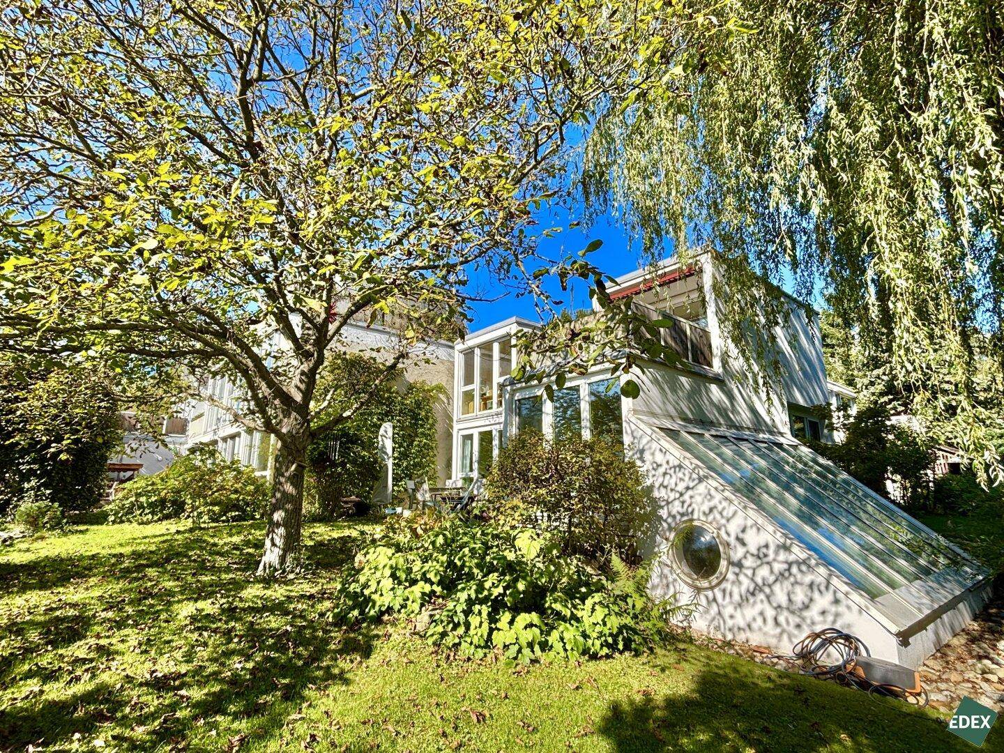 Modernes Gebäude mit großen Glasflächen und schrägem Glasdach, eingebettet in einen grünen Garten.