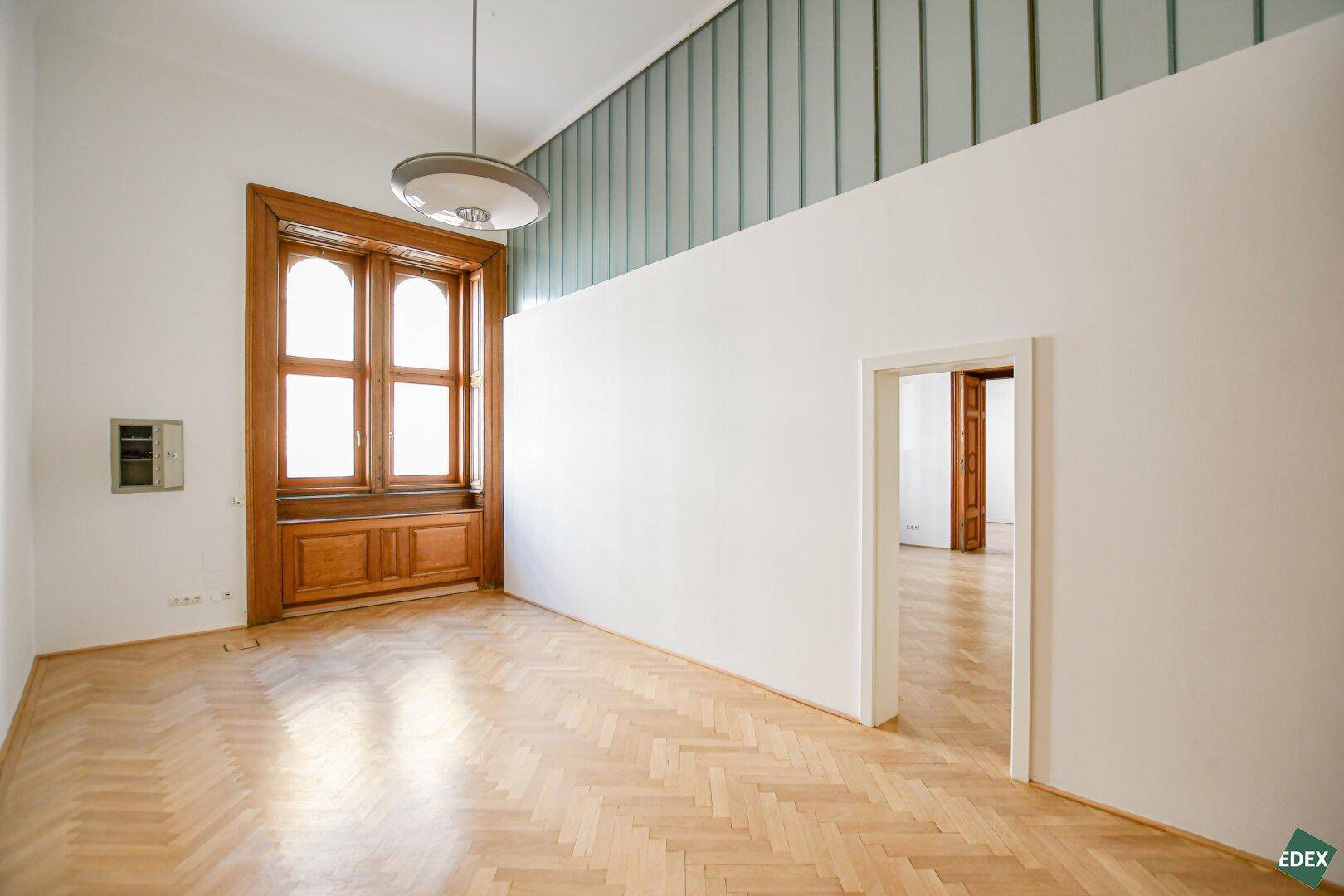 Heller Büroraum mit Fischgrätparkett, hohen Decken und einer modernen Glasabtrennung im oberen Bereich.