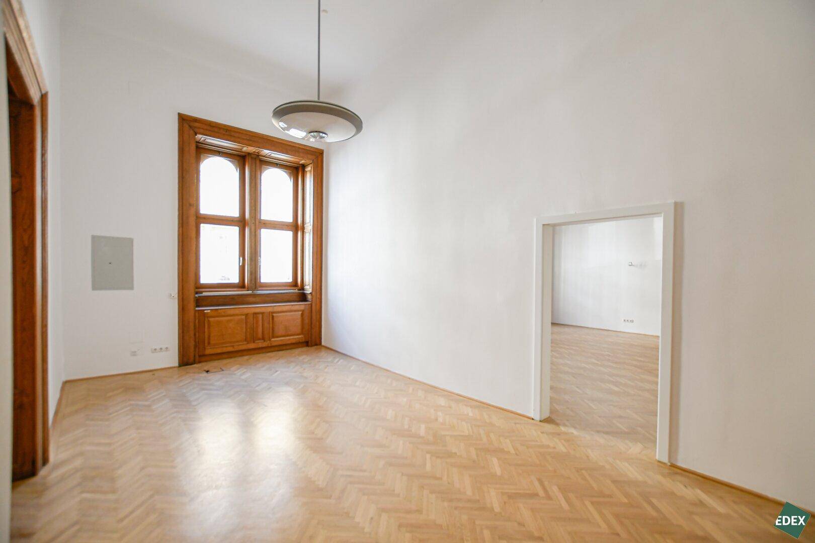 Großer, leerer Büroraum mit klassischem Fischgrätparkett, hohen Decken und einem markanten Holzfenster.